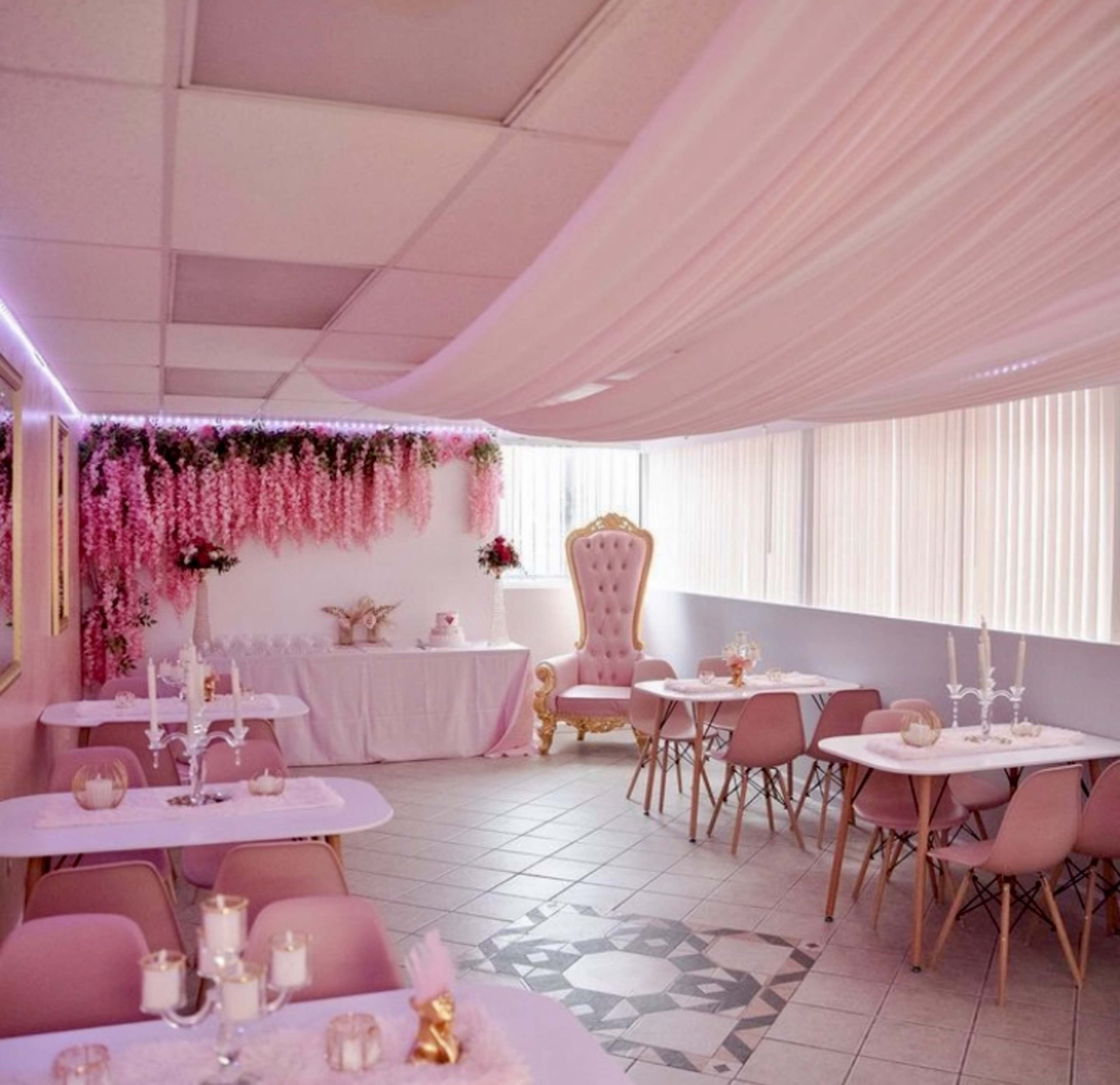 The image shows a decorated event space with pink tables and chairs, draped fabric on the ceiling, and floral arrangements on the walls.