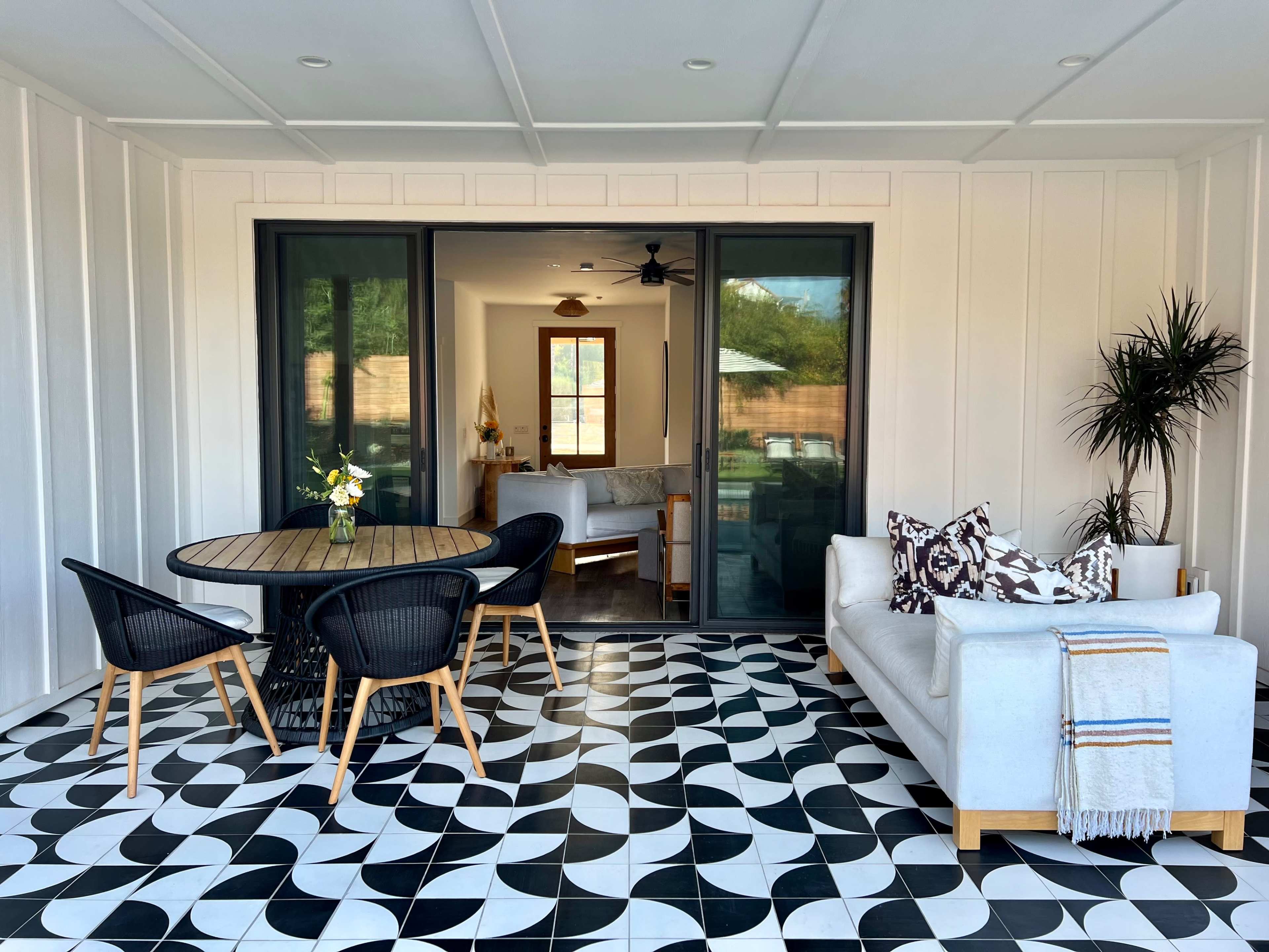 A patio features a round wooden table surrounded by black chairs and a white sofa, with a view into a room through large sliding glass doors.
