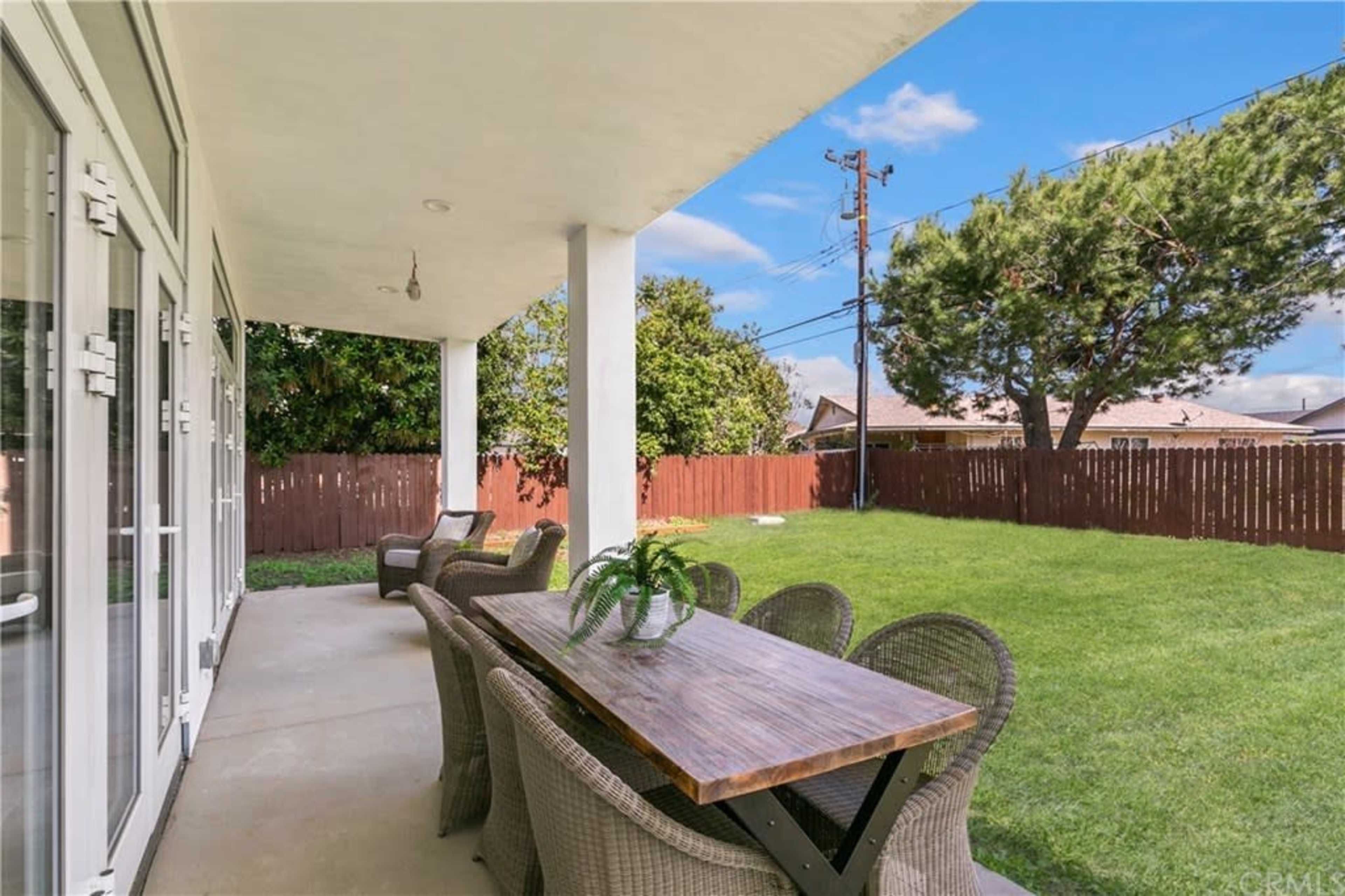 A covered patio features a long wooden table surrounded by wicker chairs, overlooking a grassy yard with a wooden fence.