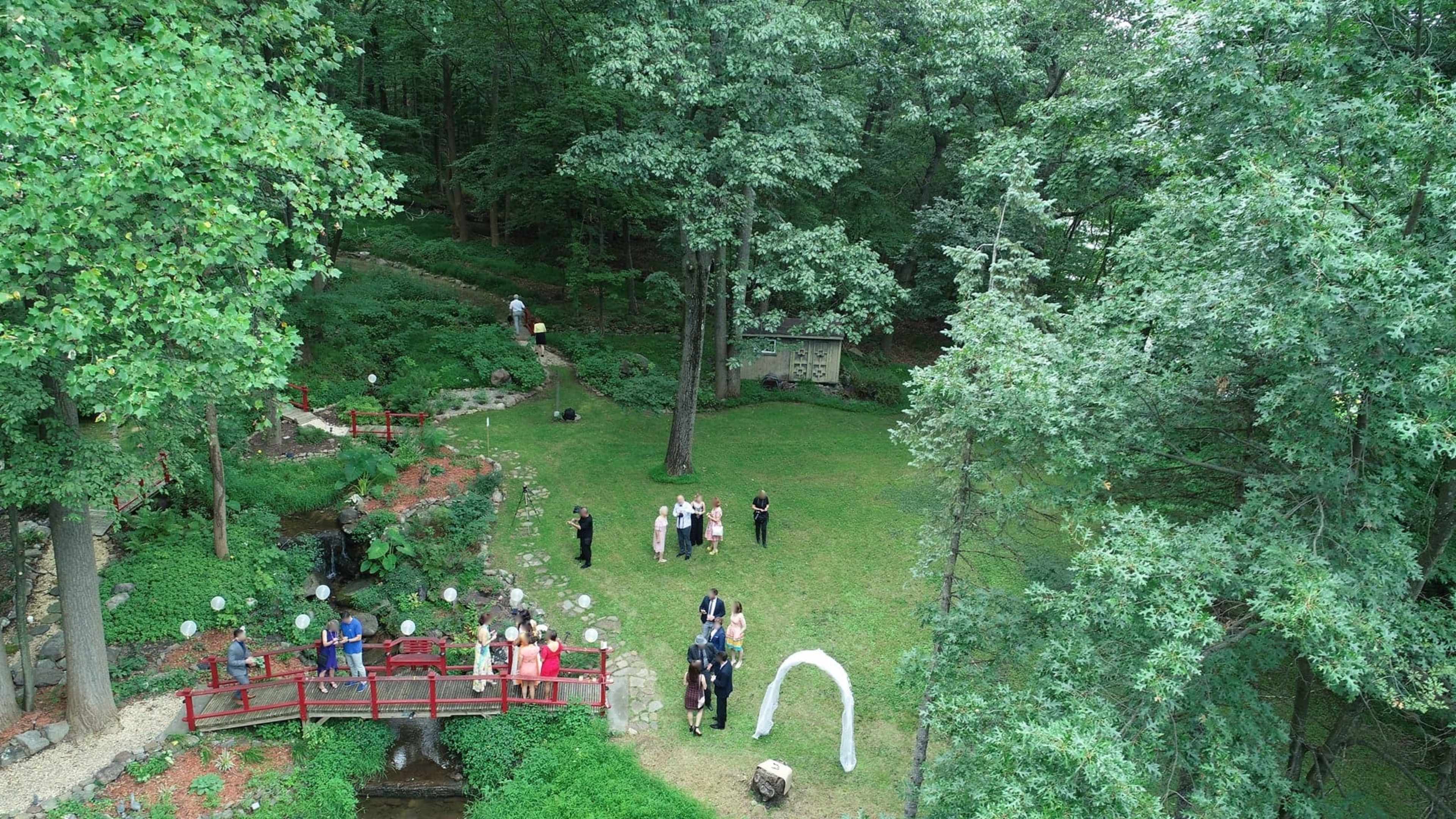A wedding ceremony is set up in a wooded area, featuring a white arch and a red bridge, with guests and participants gathered on a grassy lawn.