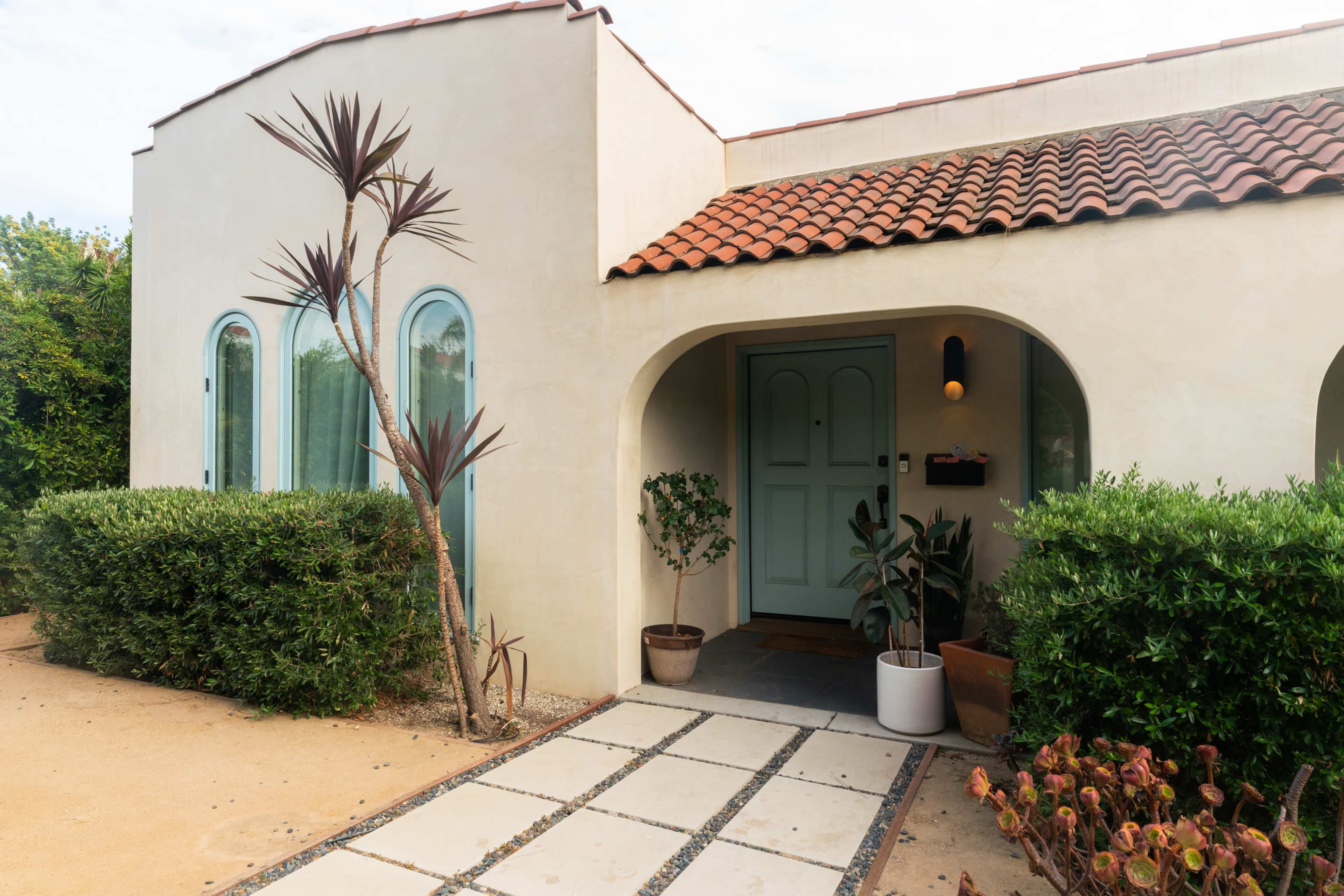 A single-story house features a rounded entrance with a light green door, surrounded by decorative plants and a pathway of square tiles leading to the front.