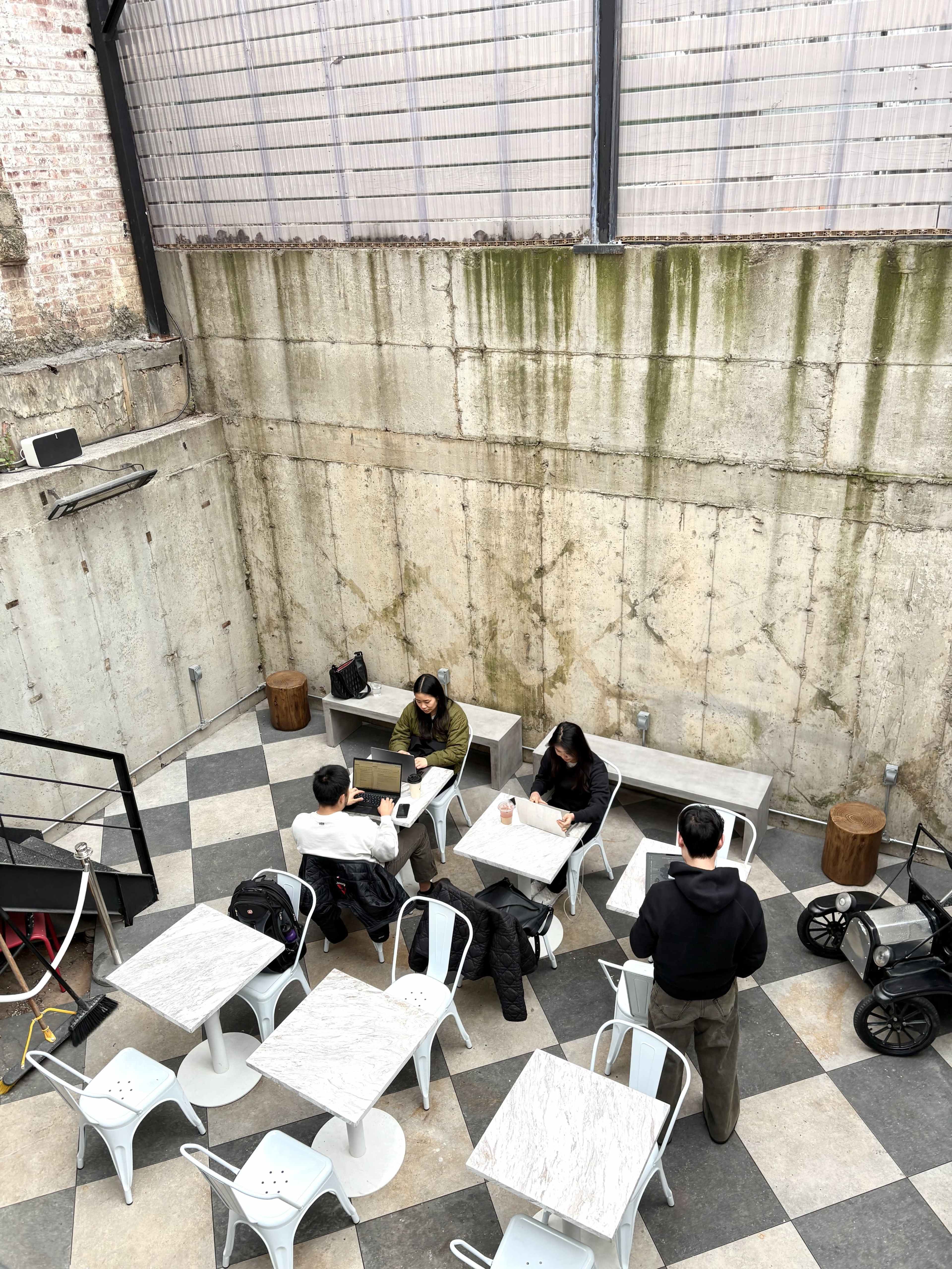 The image shows a small outdoor cafe with several people seated at tables, working on laptops and writing, against a backdrop of bare concrete walls and a checkered floor.