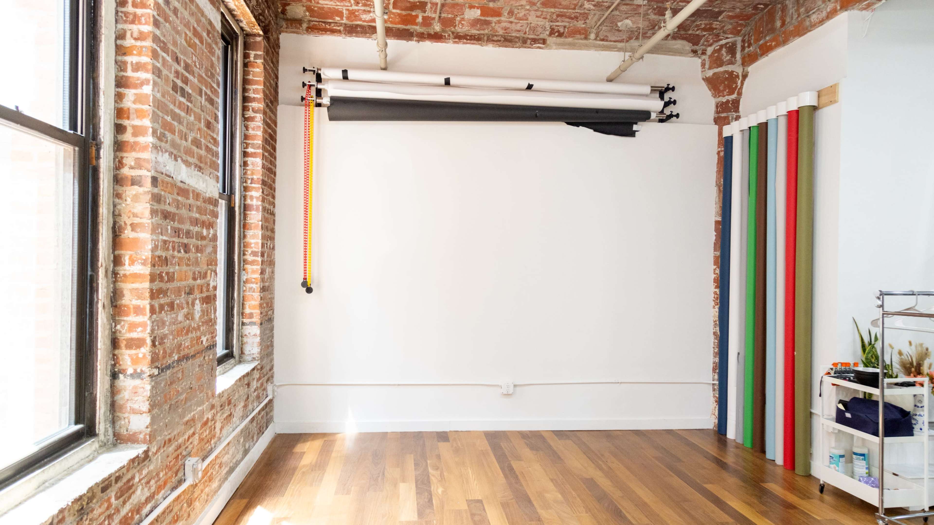 A studio space with exposed brick walls, hardwood flooring, and a collection of backdrop rolls on the right side.