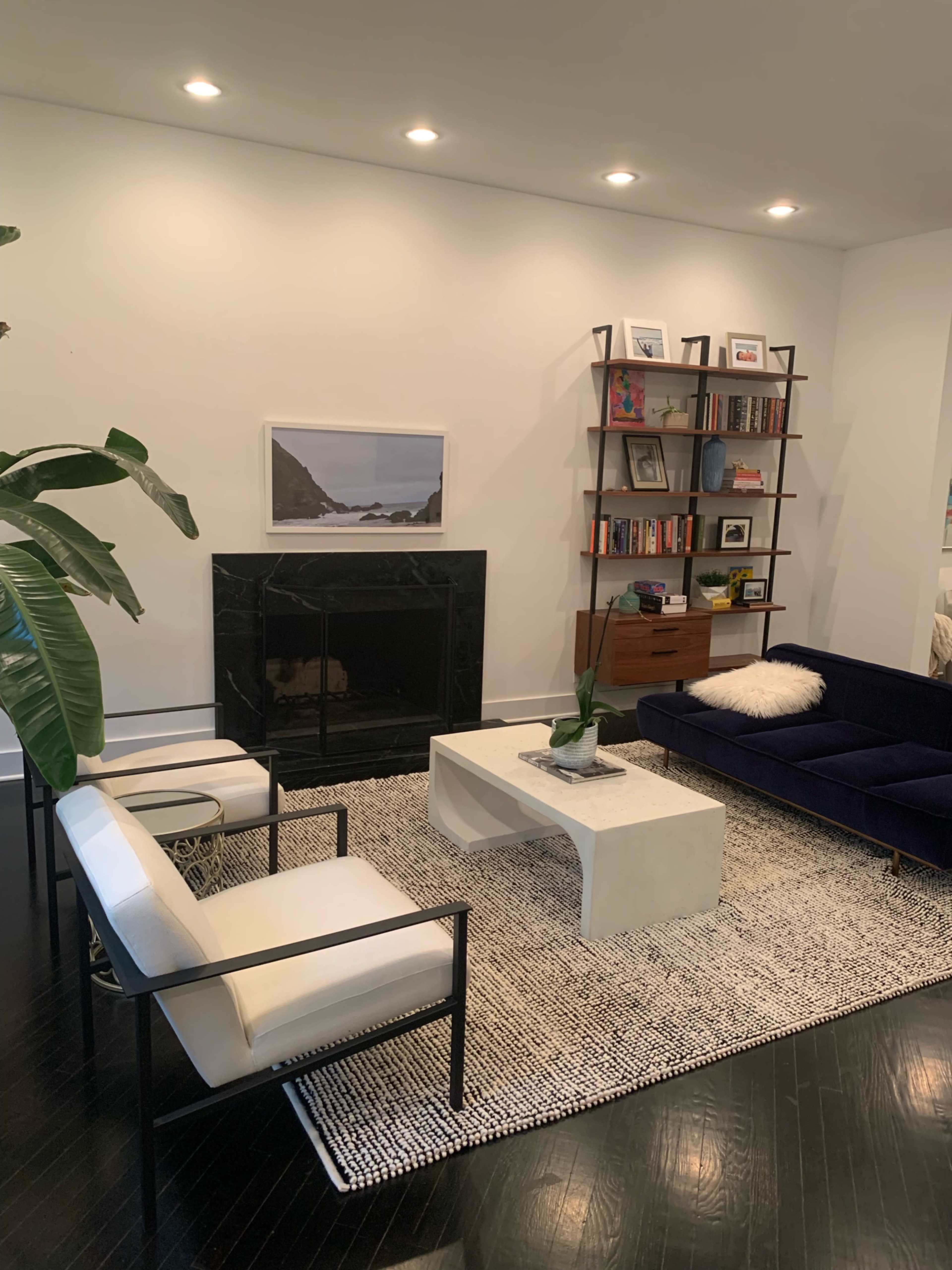 A modern living room featuring a dark blue sofa, a white coffee table, a black fireplace, a bookshelf filled with books, and plants in a well-organized layout.