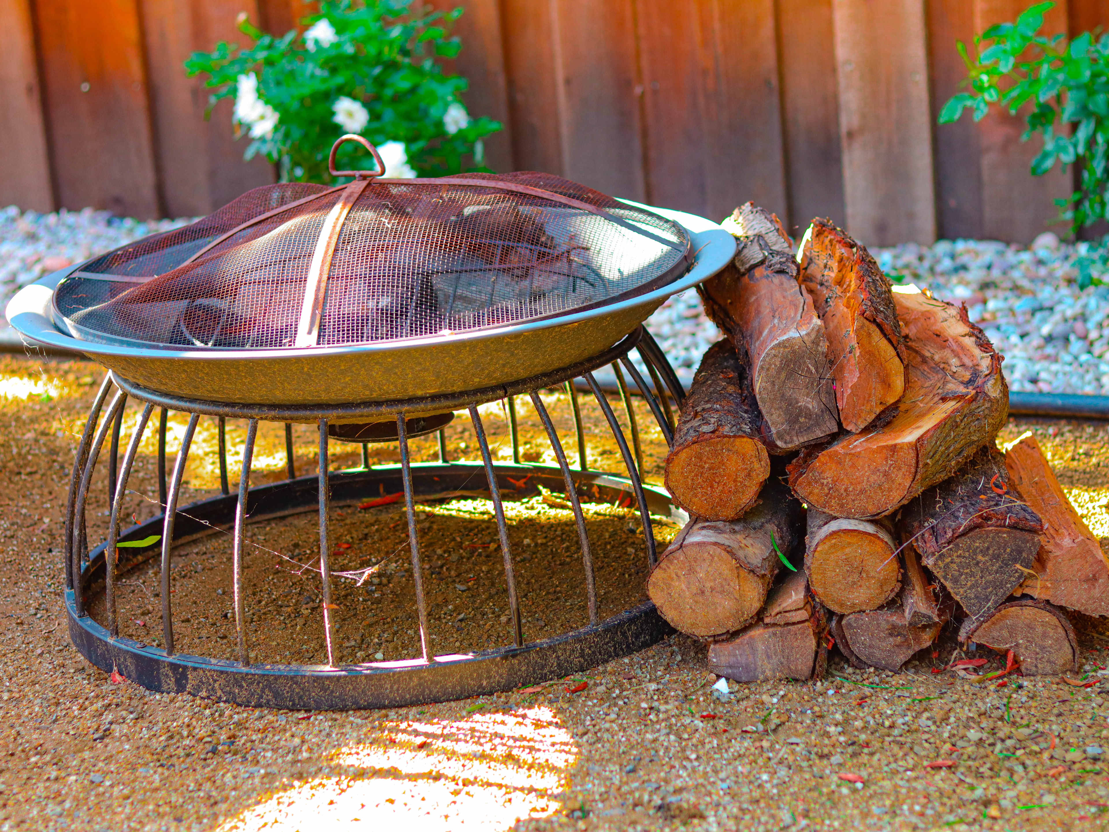 A fire pit with a mesh cover sits next to a stack of neatly arranged firewood on a gravel surface.