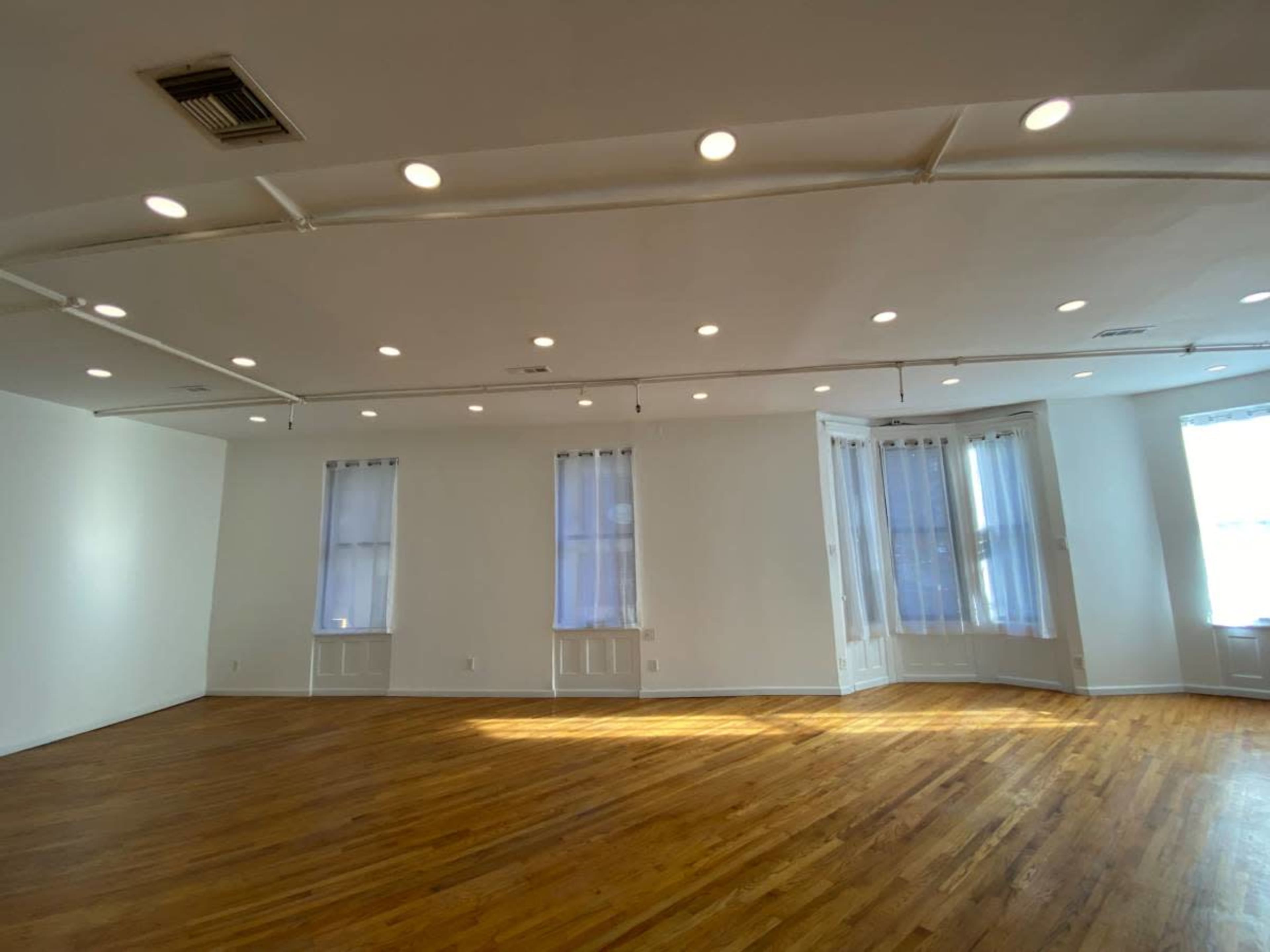 The image shows a spacious, empty room with hardwood floors, white walls, and multiple windows allowing natural light to enter.