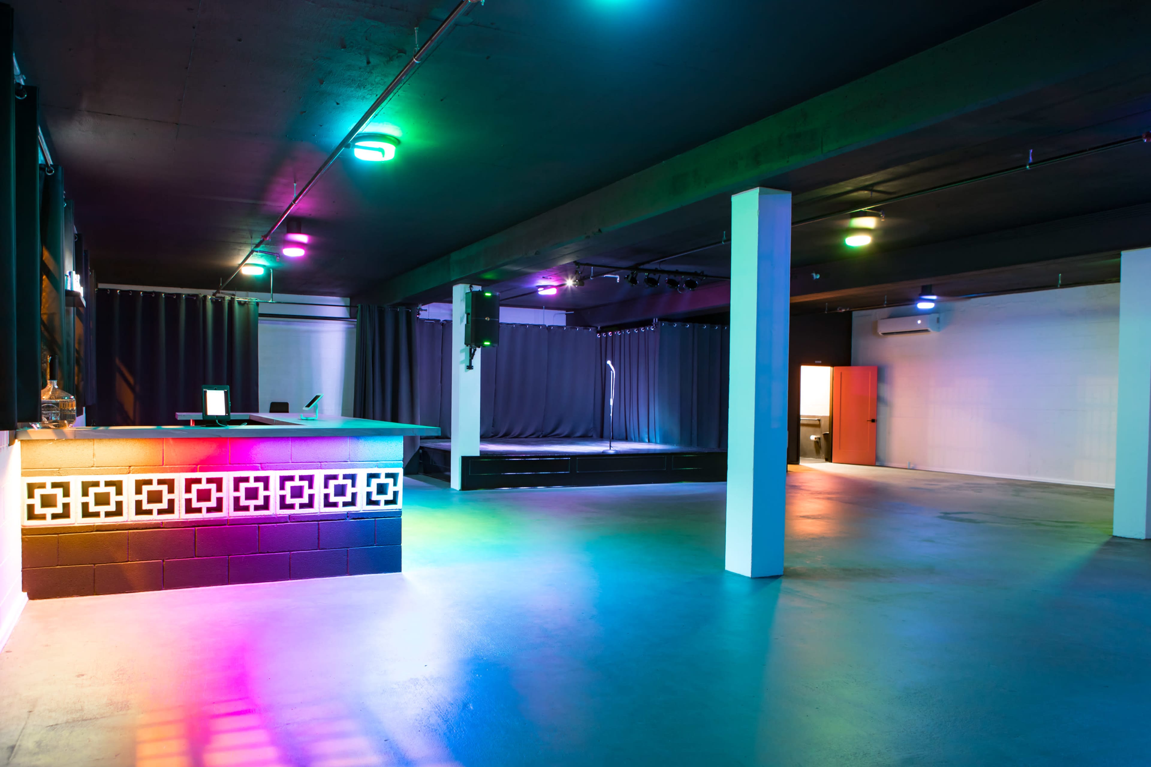 A dimly lit event space with a bar area, stage, and colorful lighting.