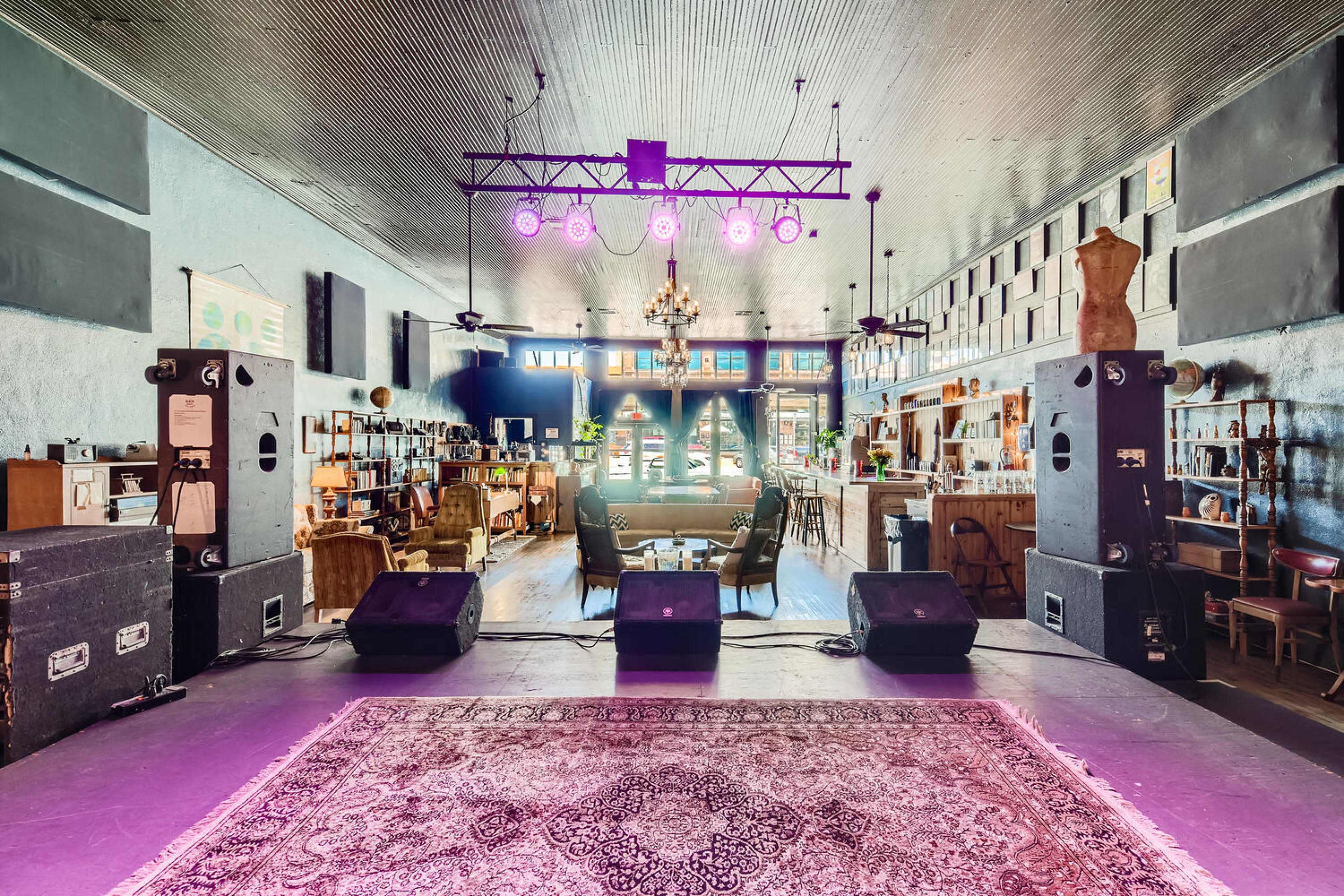 The image shows a performance stage with speakers and microphones in a venue featuring a bar and seating area, illuminated by overhead lights.