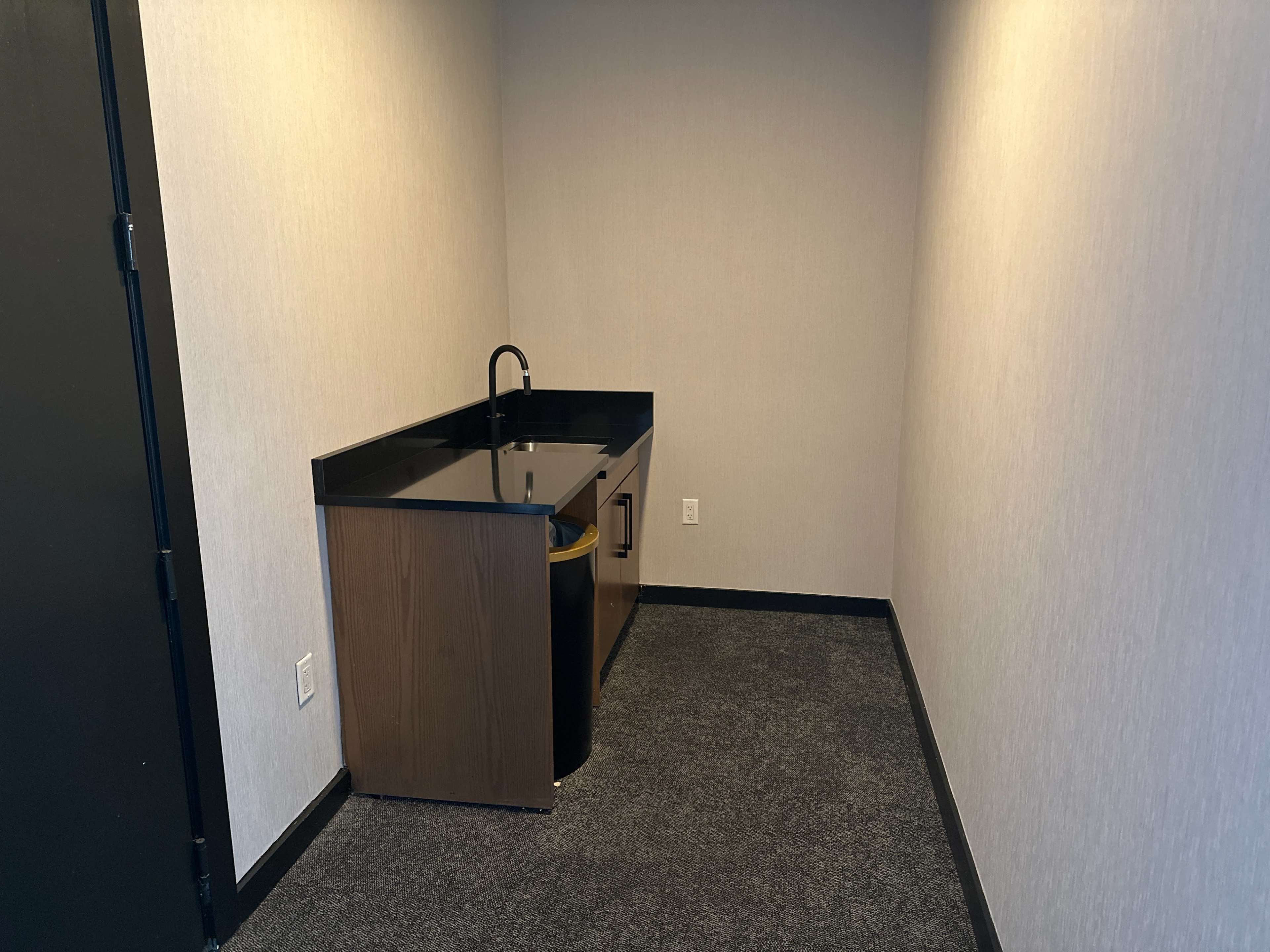 The image shows a small, empty room with a countertop and sink against one wall, featuring black surfaces and gray carpet.