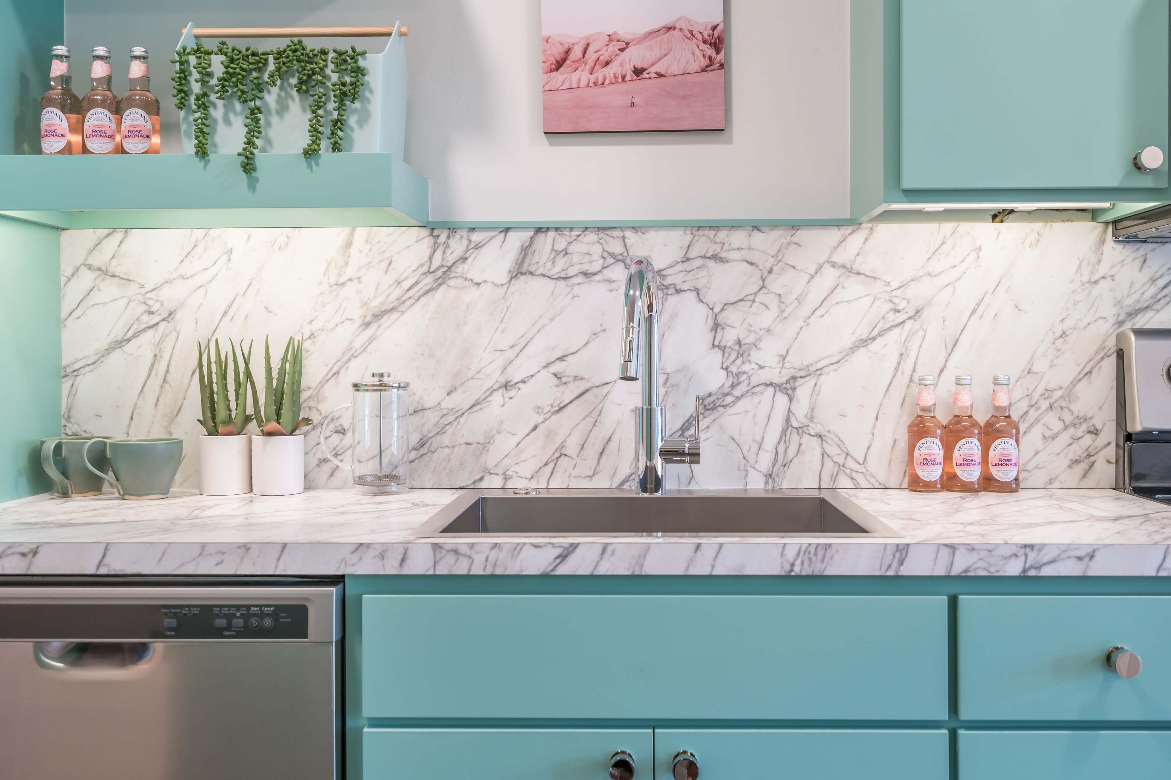 A modern kitchen with teal cabinets, a marble countertop, and decorative elements including plants and beverages on the counter.