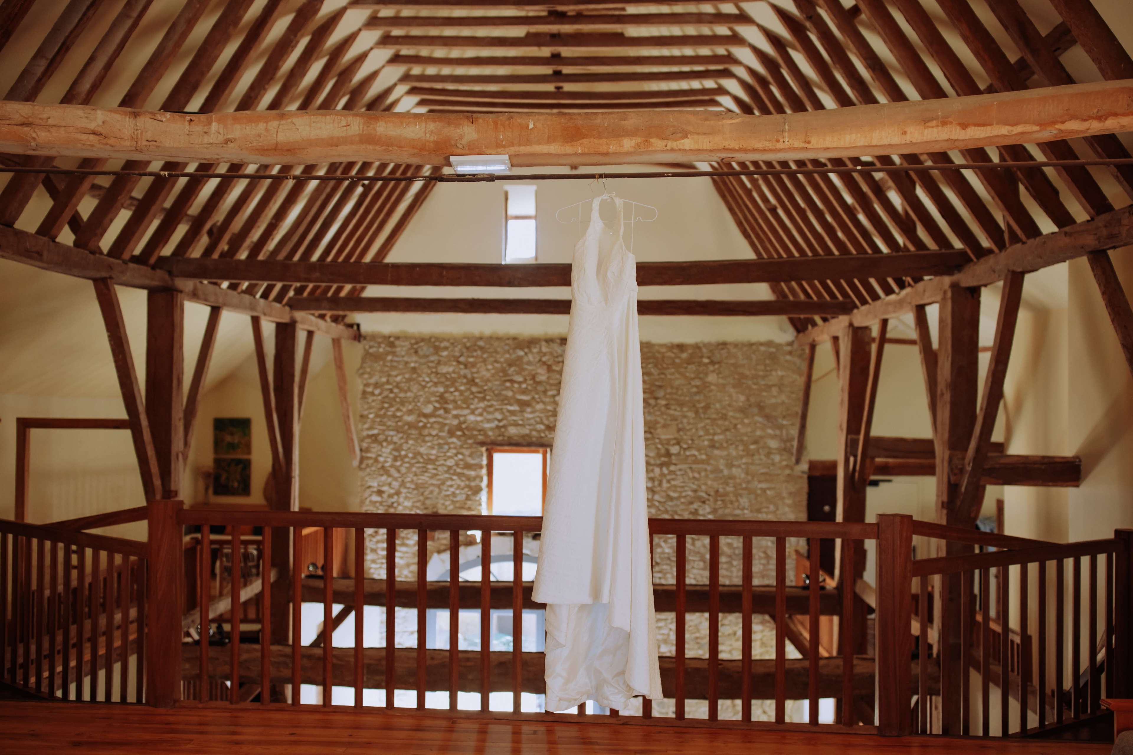 A white wedding dress is hanging on a wooden railing in a spacious, rustic room with exposed beams and stone walls.
