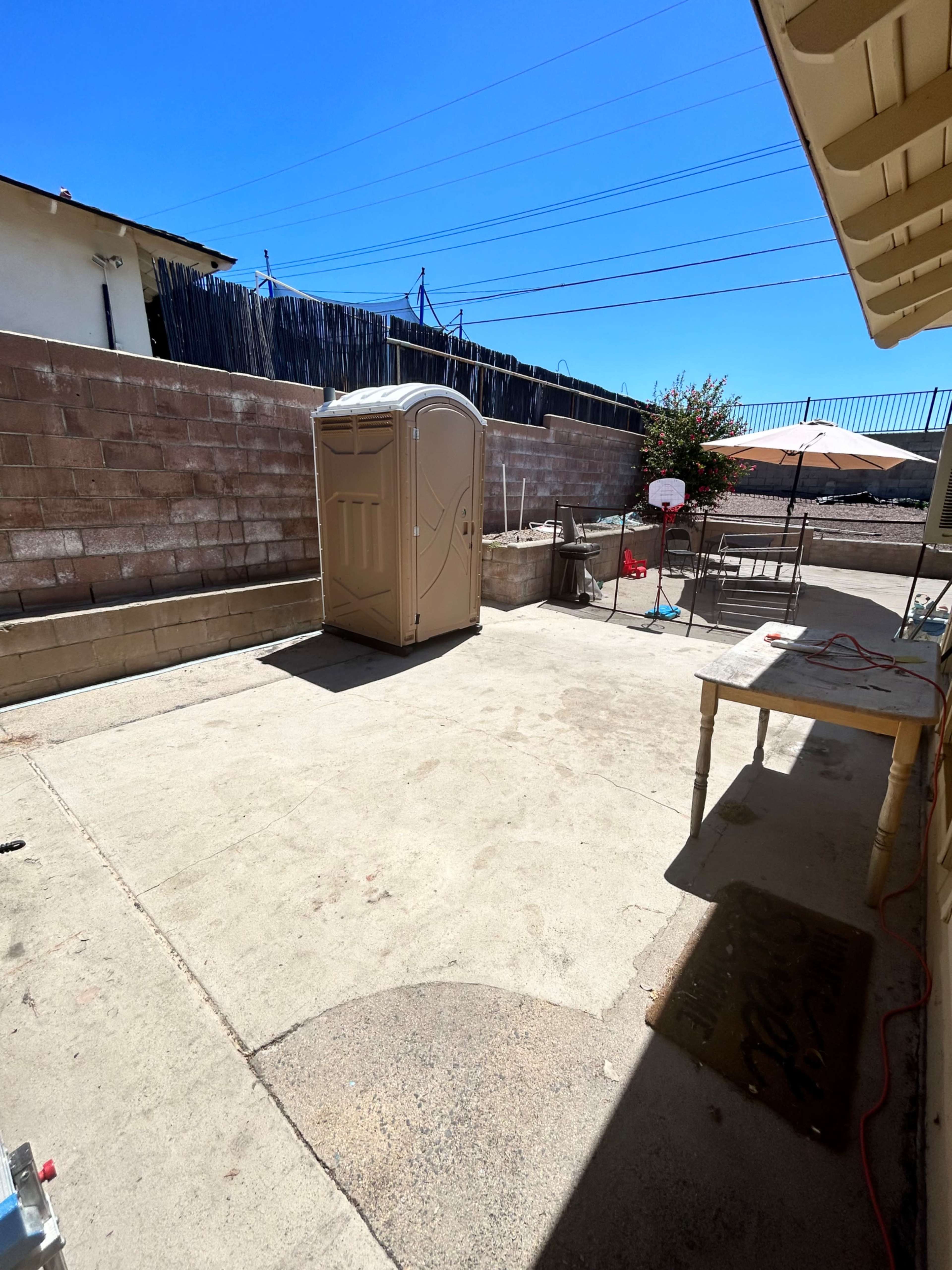 A concrete outdoor area with a portable restroom, a table, and a fence in the background under a clear blue sky.