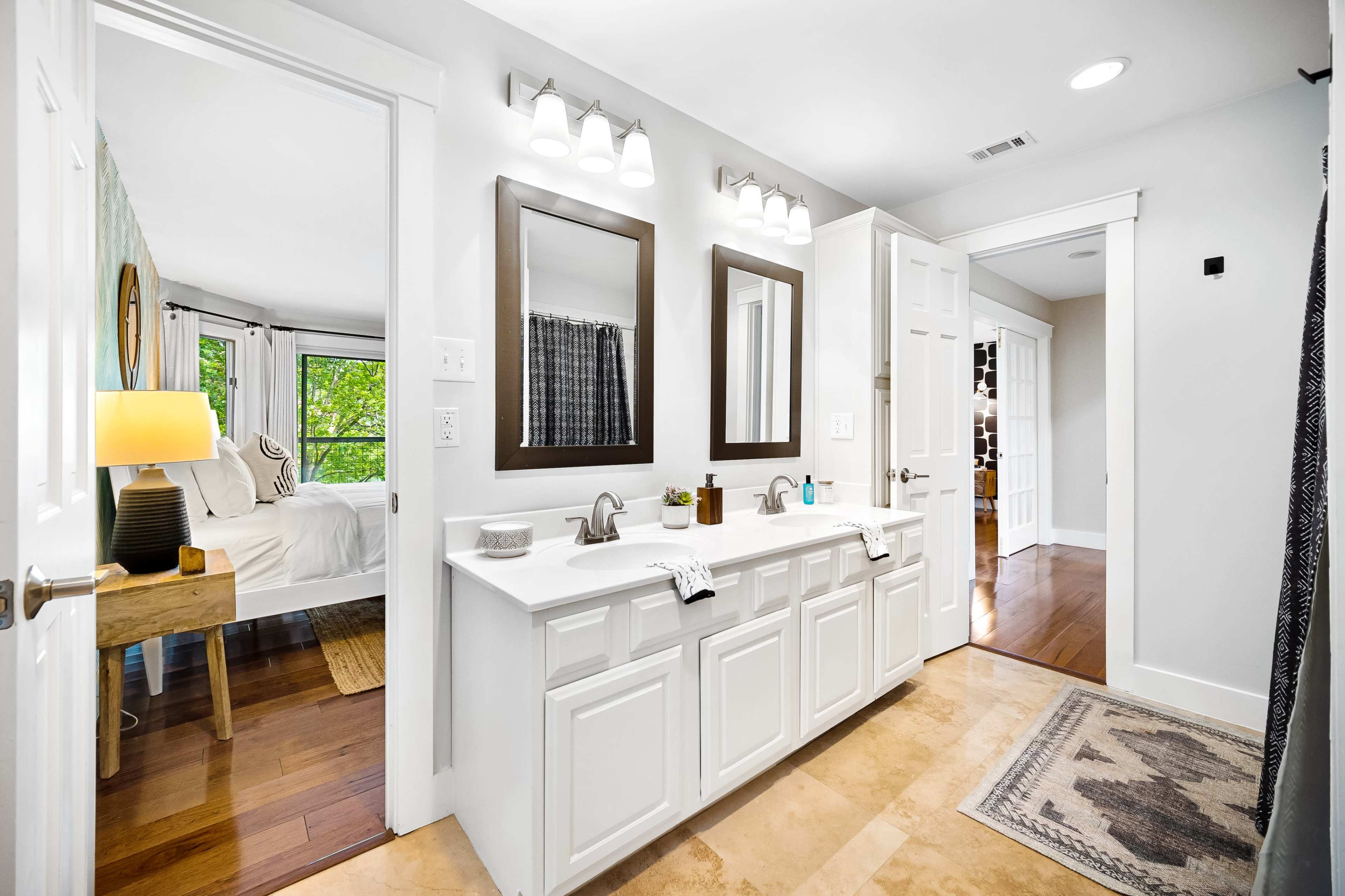 The image shows a well-lit bathroom featuring double sinks with mirrors, a wooden table lamp, and a hallway leading to other rooms.