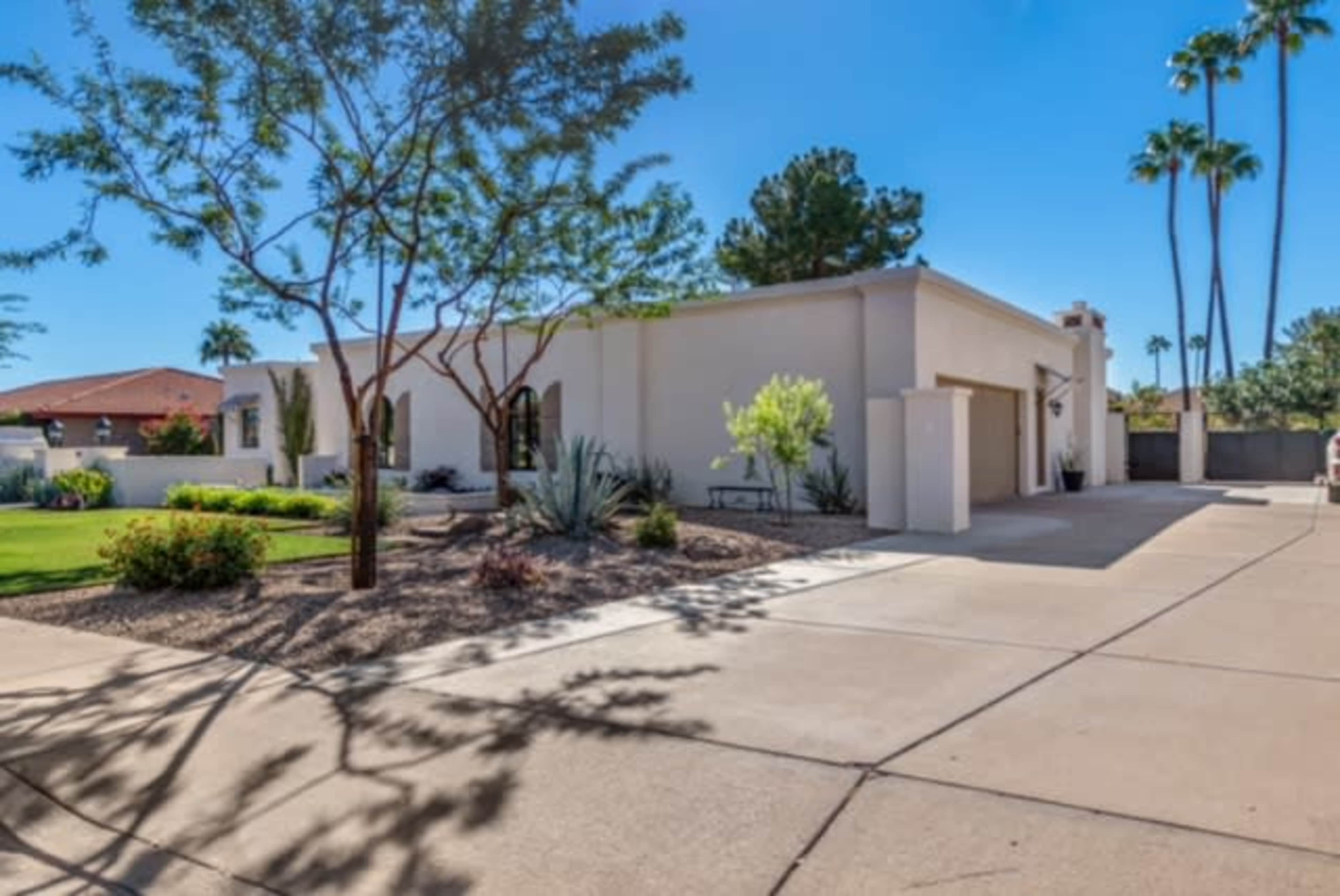 A single-story, modern house is surrounded by desert landscaping and palm trees, with a driveway leading to the garage.