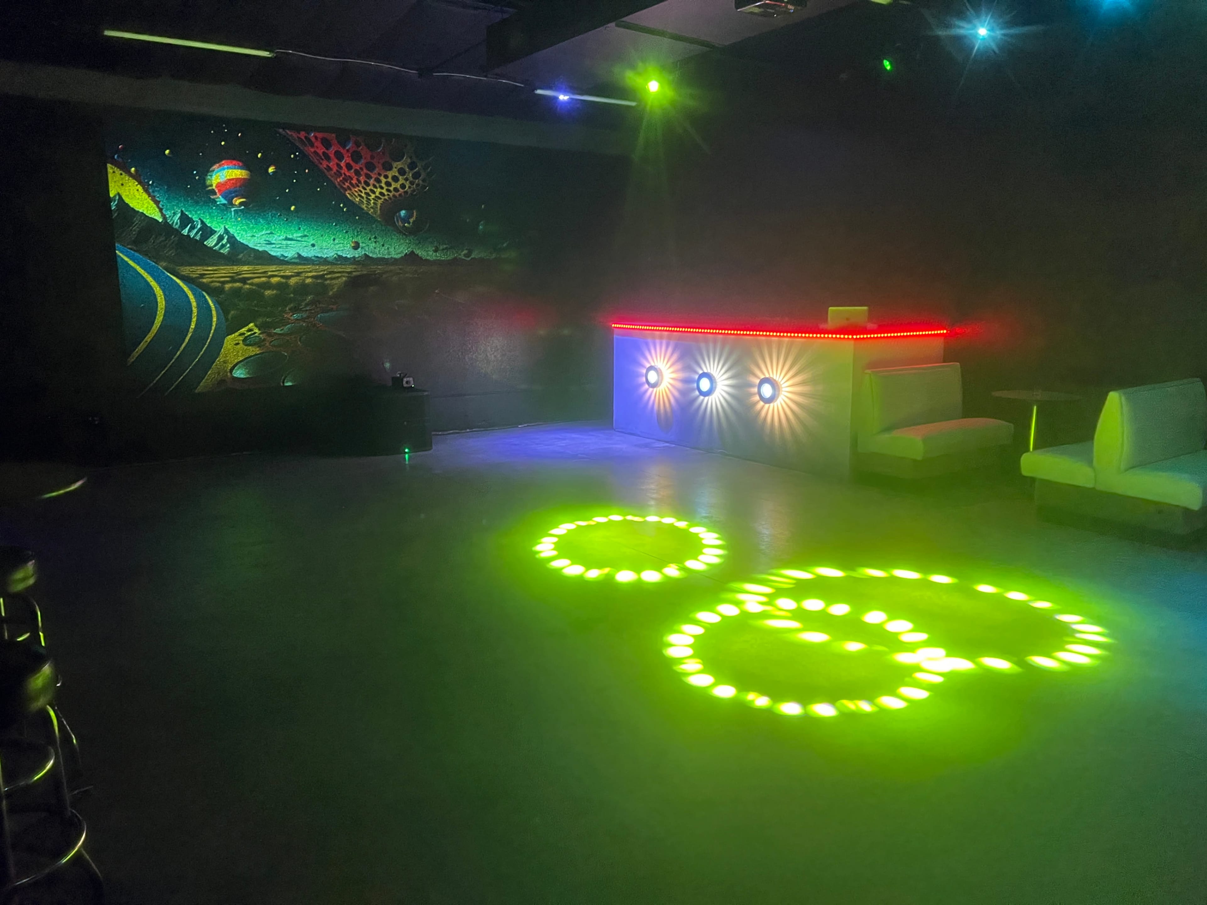 The image shows a dimly lit room with a colorful mural on one wall, a bar with illuminated edges, and green lighting casting circular patterns on the floor.