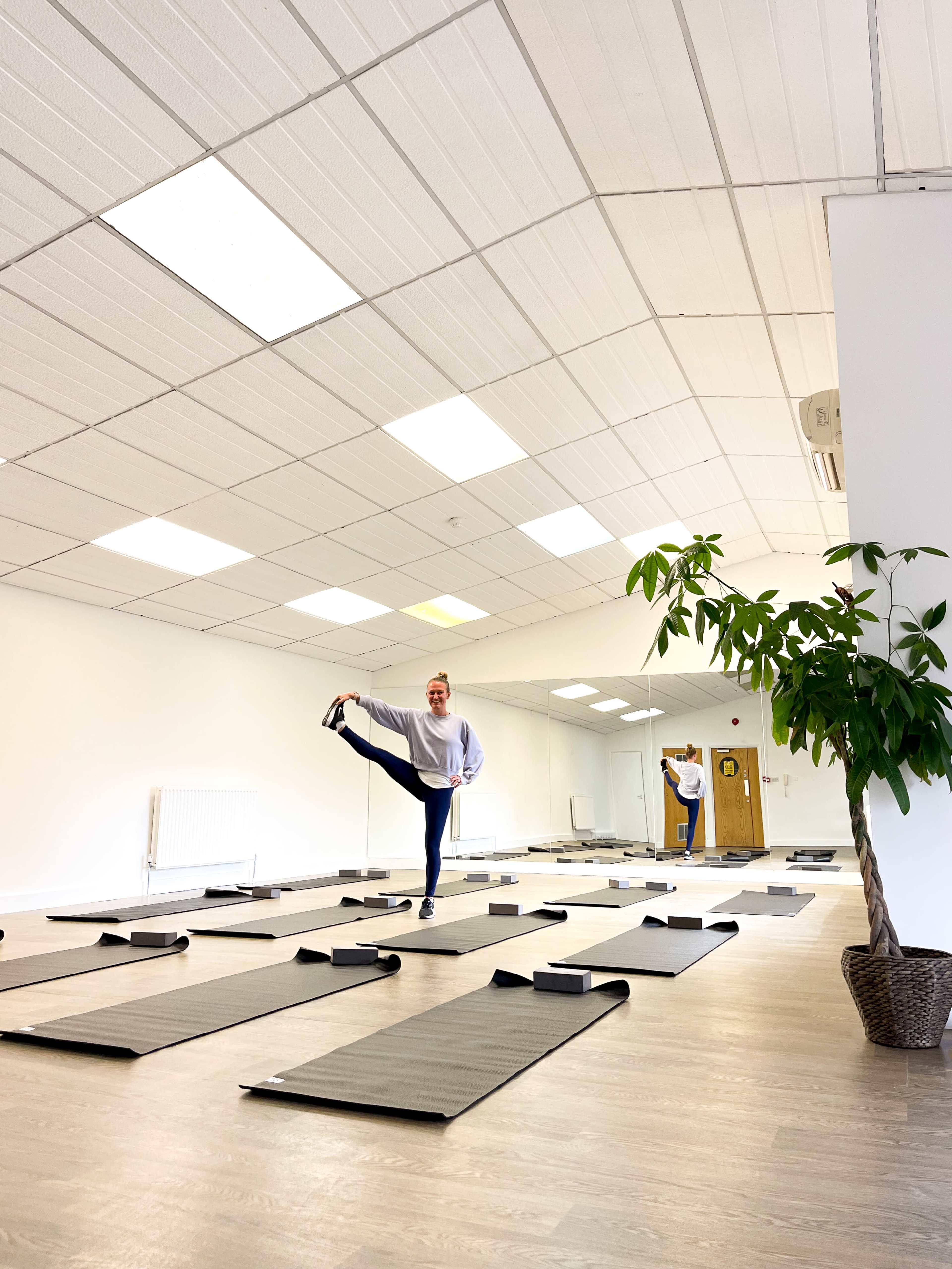 A woman performs a yoga pose on one leg in a bright studio with several yoga mats arranged on the floor.
