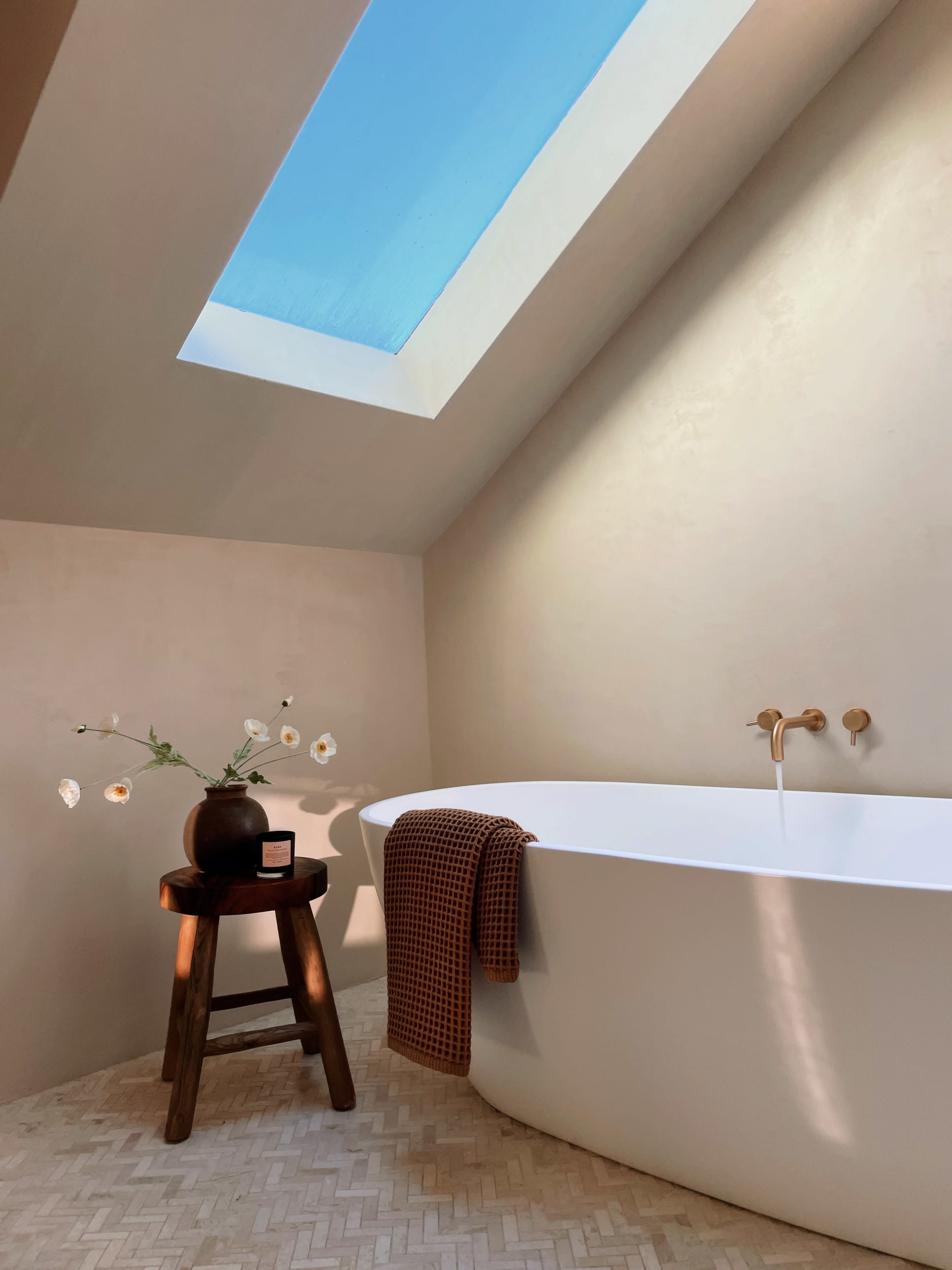 A modern bathroom features a freestanding white bathtub, a wooden stool with a vase of flowers, and a skylight that illuminates the space.