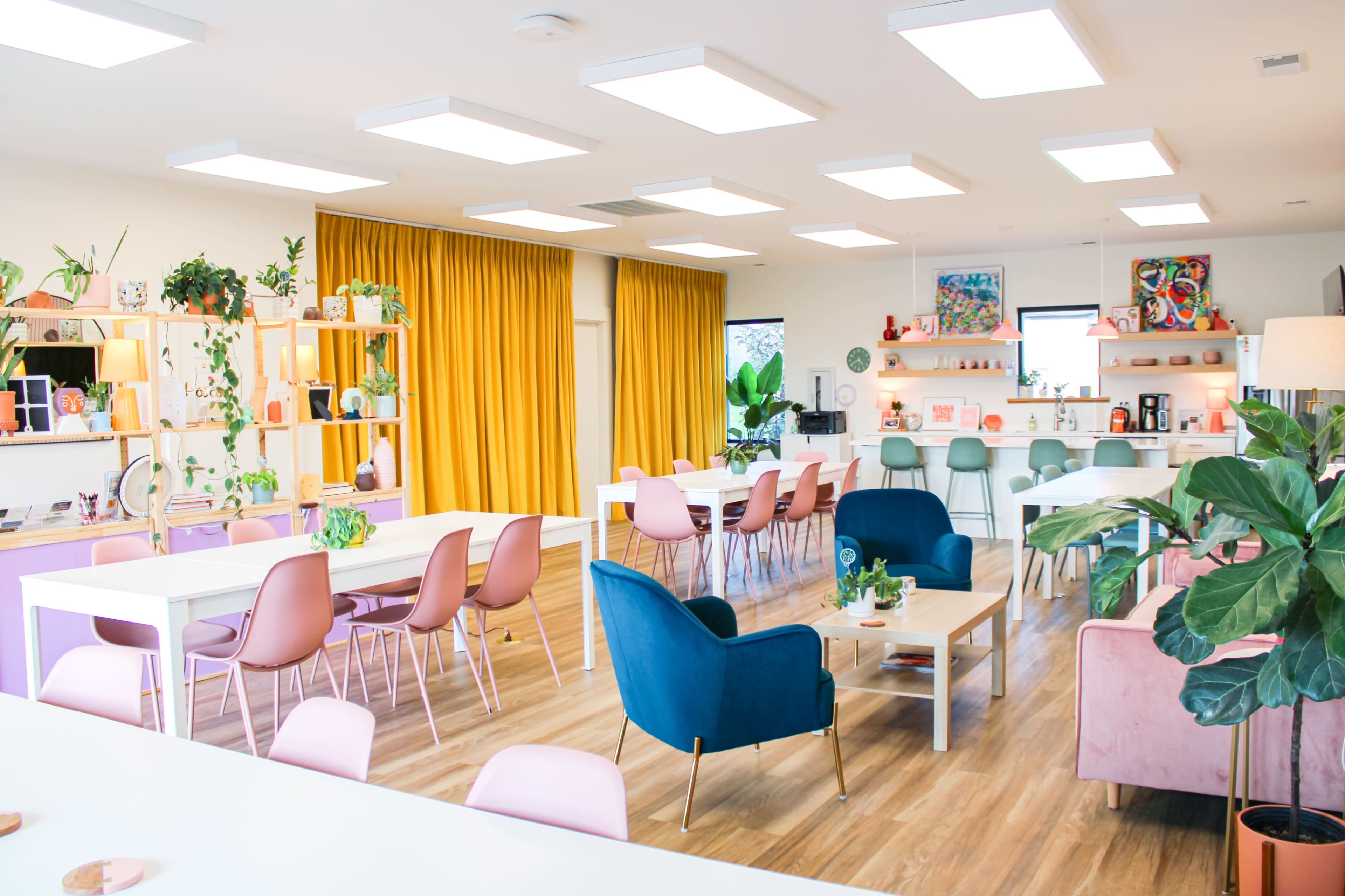 The image shows a modern office space with multiple work tables, colorful chairs, a kitchenette area, and large windows with yellow curtains.