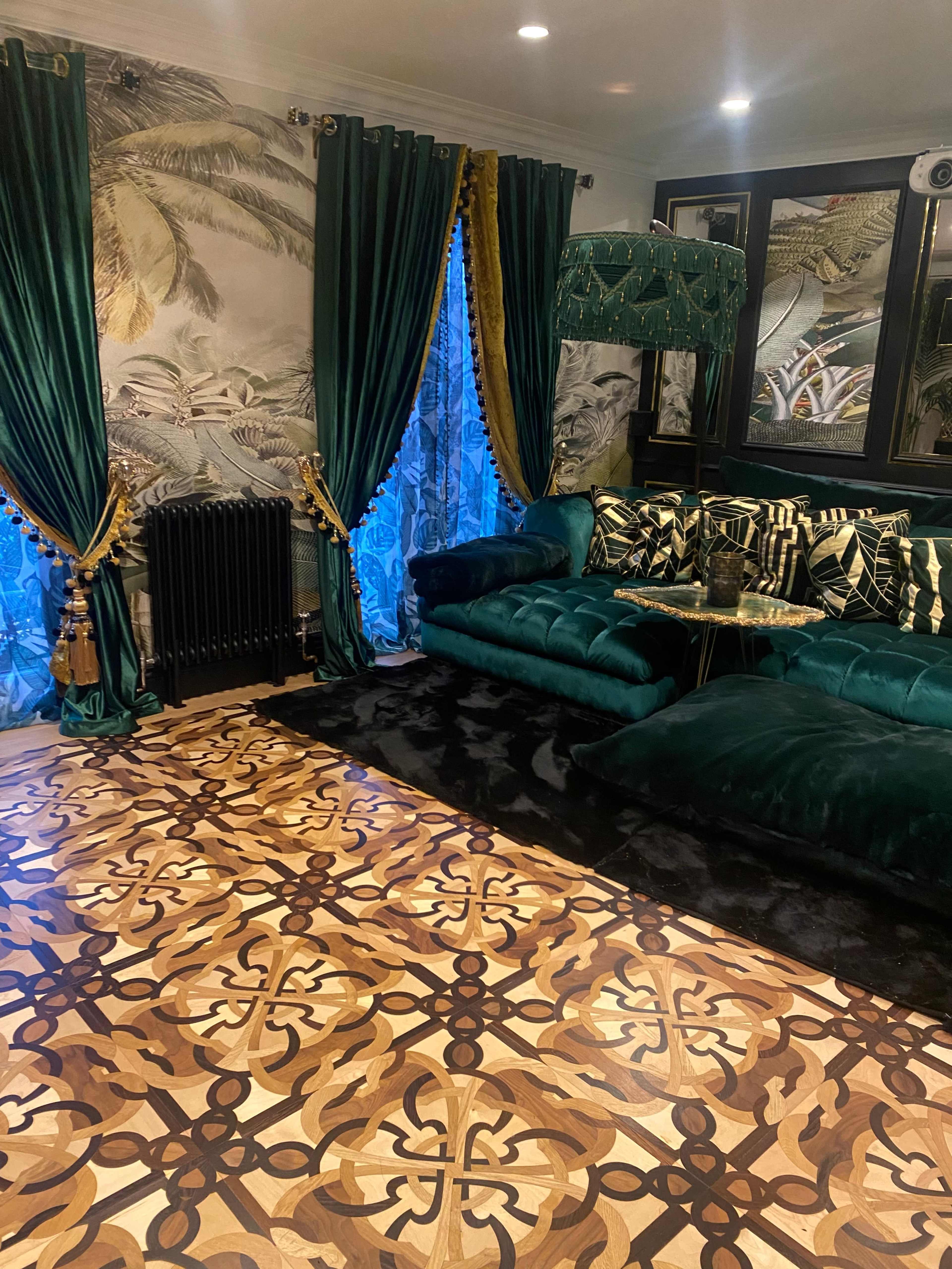 A living room featuring a dark green velvet sectional sofa, intricate wooden flooring, and patterned wallpaper with botanical designs.