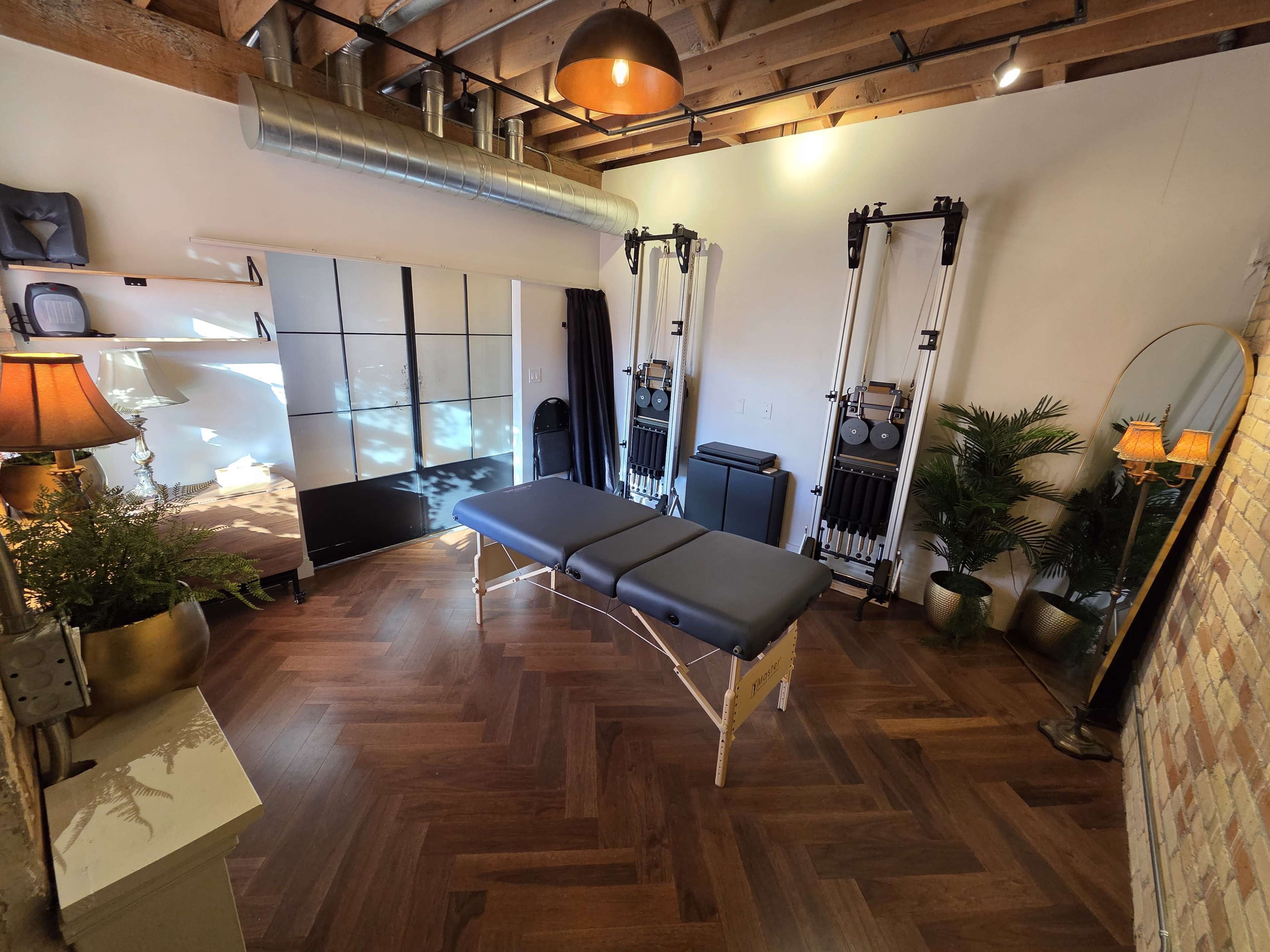 The image shows a well-appointed massage therapy room featuring a massage table, exercise equipment, a large mirror, plants, and warm lighting.