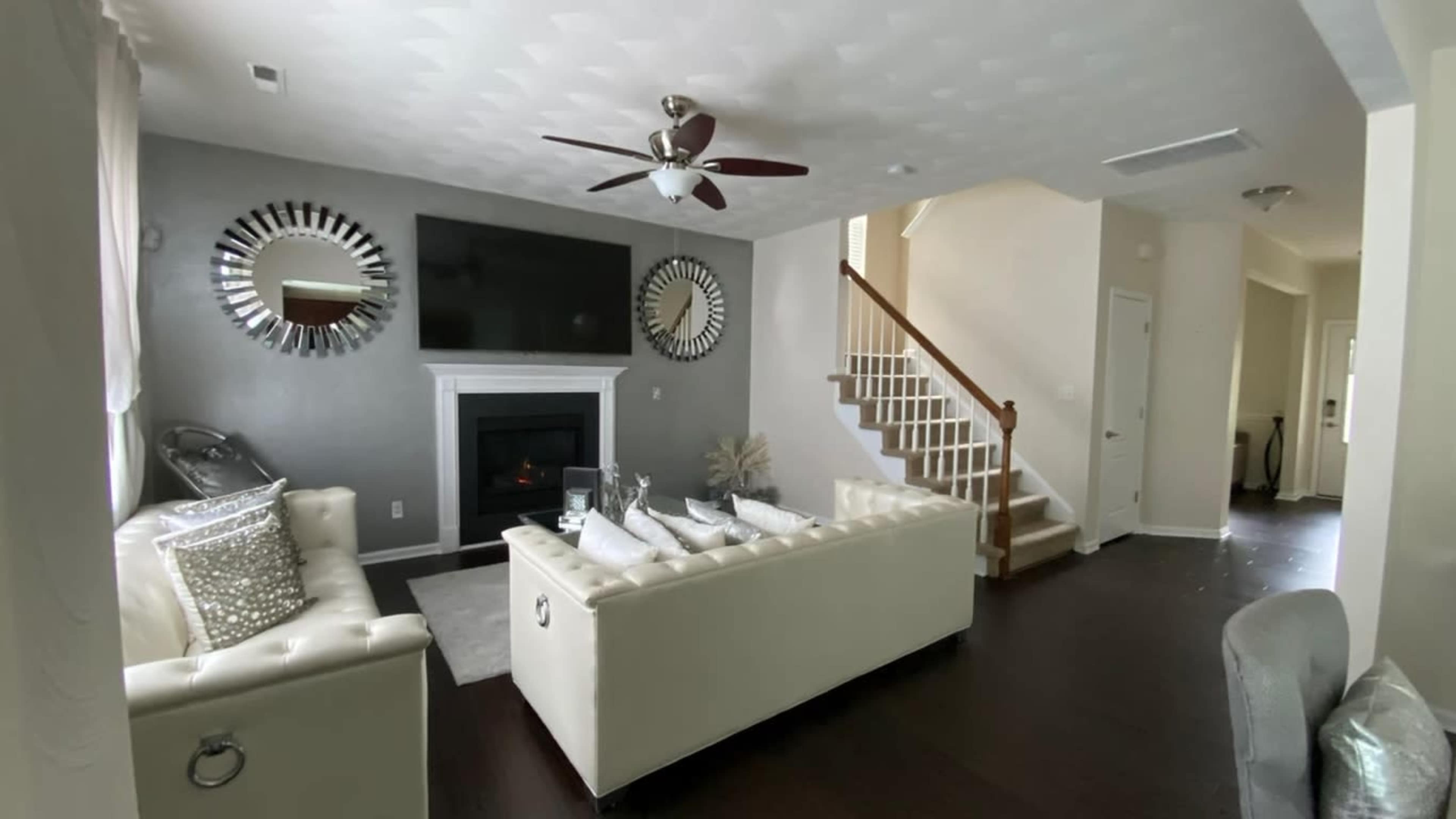 A modern living room features a white tufted sofa, a large flat-screen TV above a fireplace, and a staircase leading to an upper level.