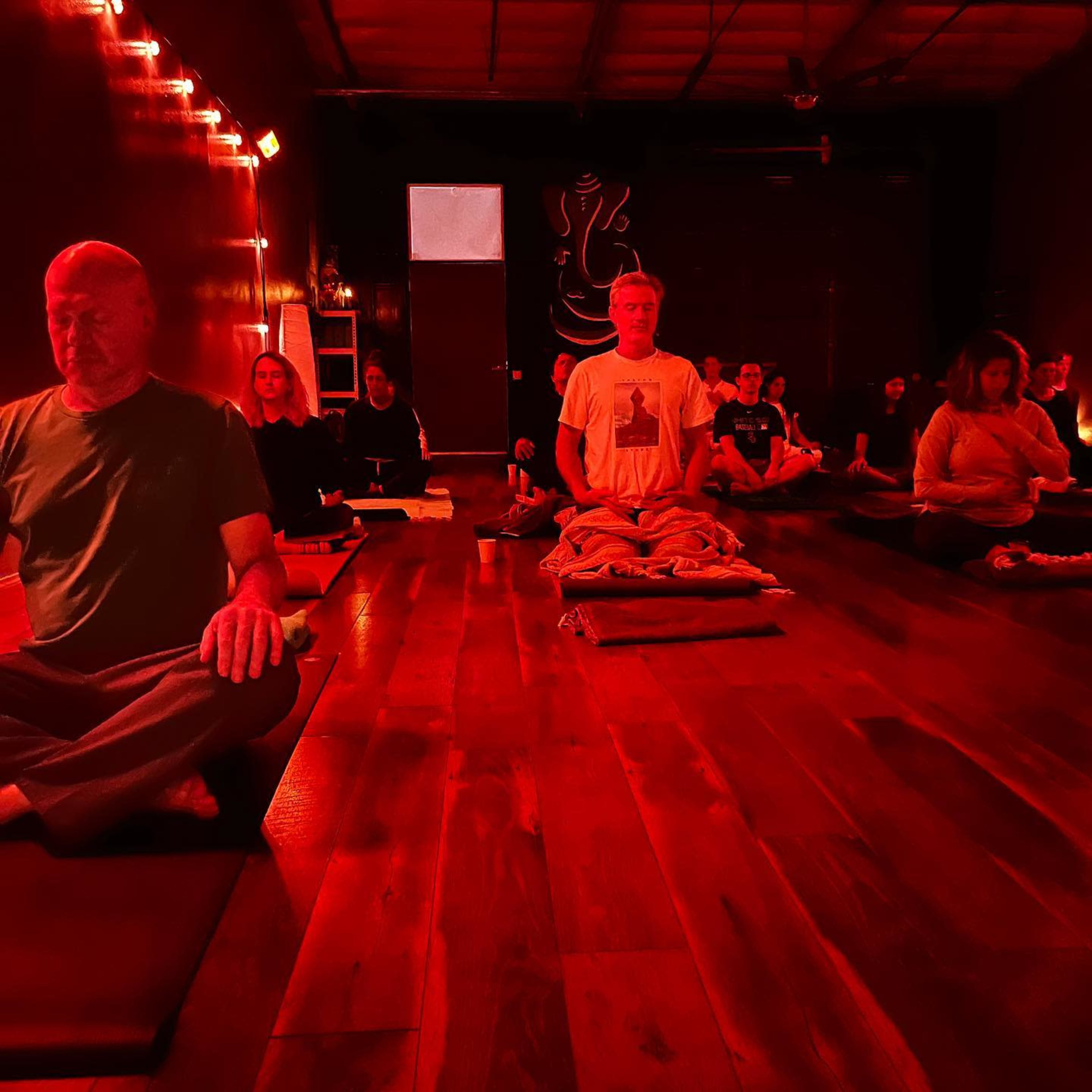 A group of individuals are seated on yoga mats in a dimly lit room, illuminated by red lights, engaged in meditation.