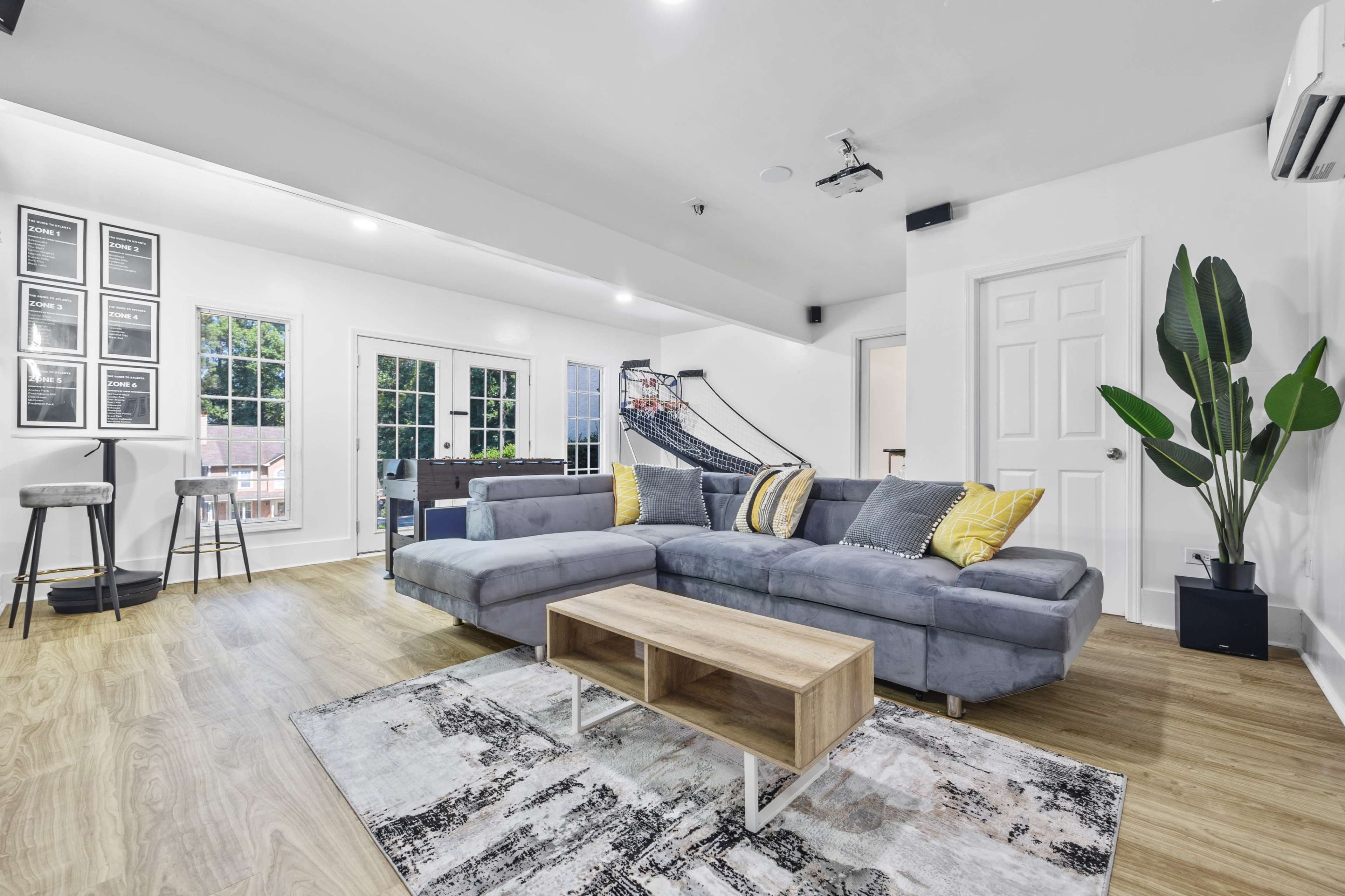 The image shows a modern living room with a gray sectional sofa, a wooden coffee table, and large windows that provide ample natural light.