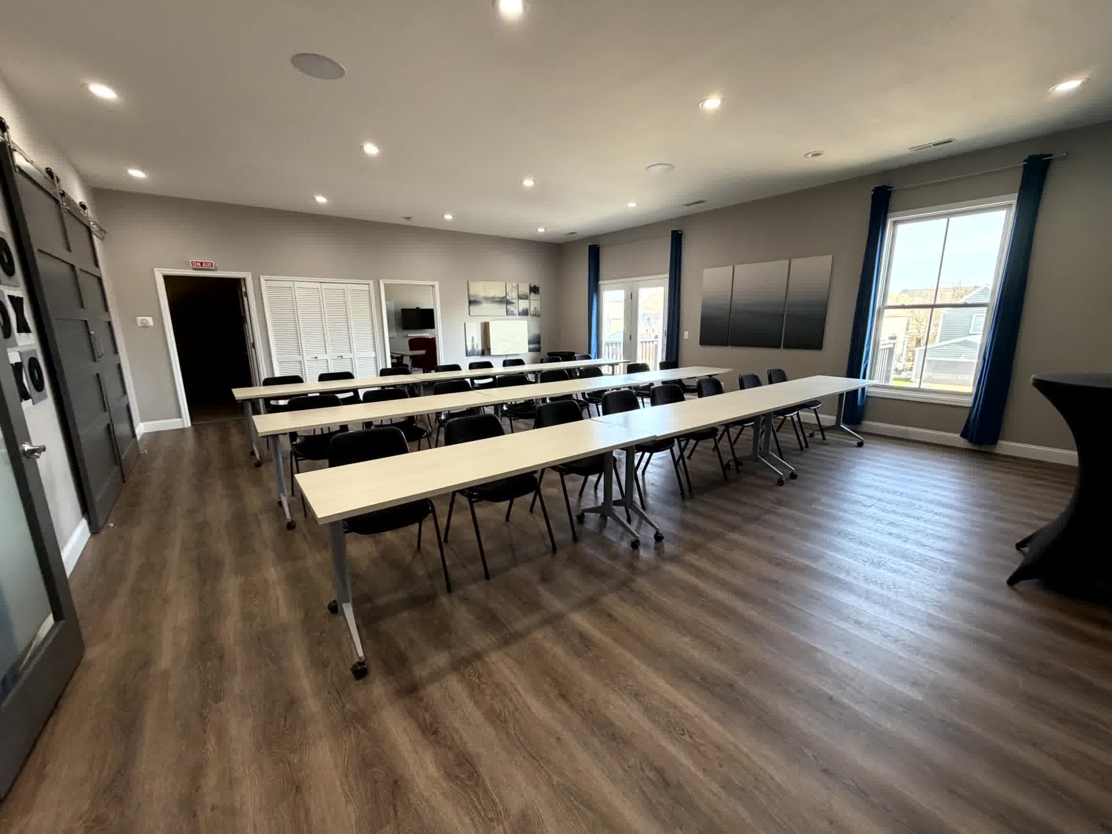 The image shows a well-lit conference room with several long tables arranged in rows, black chairs, and large windows letting in natural light.