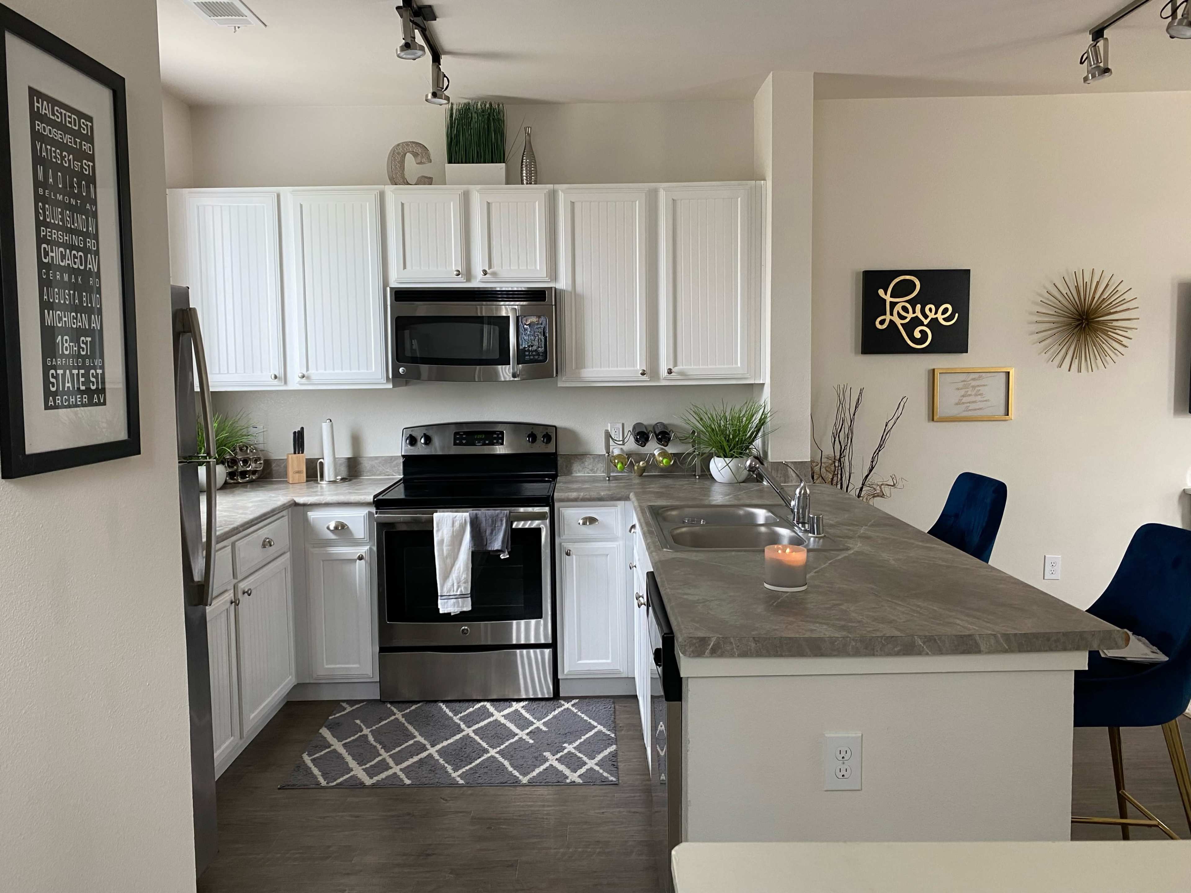 A modern kitchen with white cabinetry, stainless steel appliances, and a gray countertop, featuring a small island with blue bar stools.