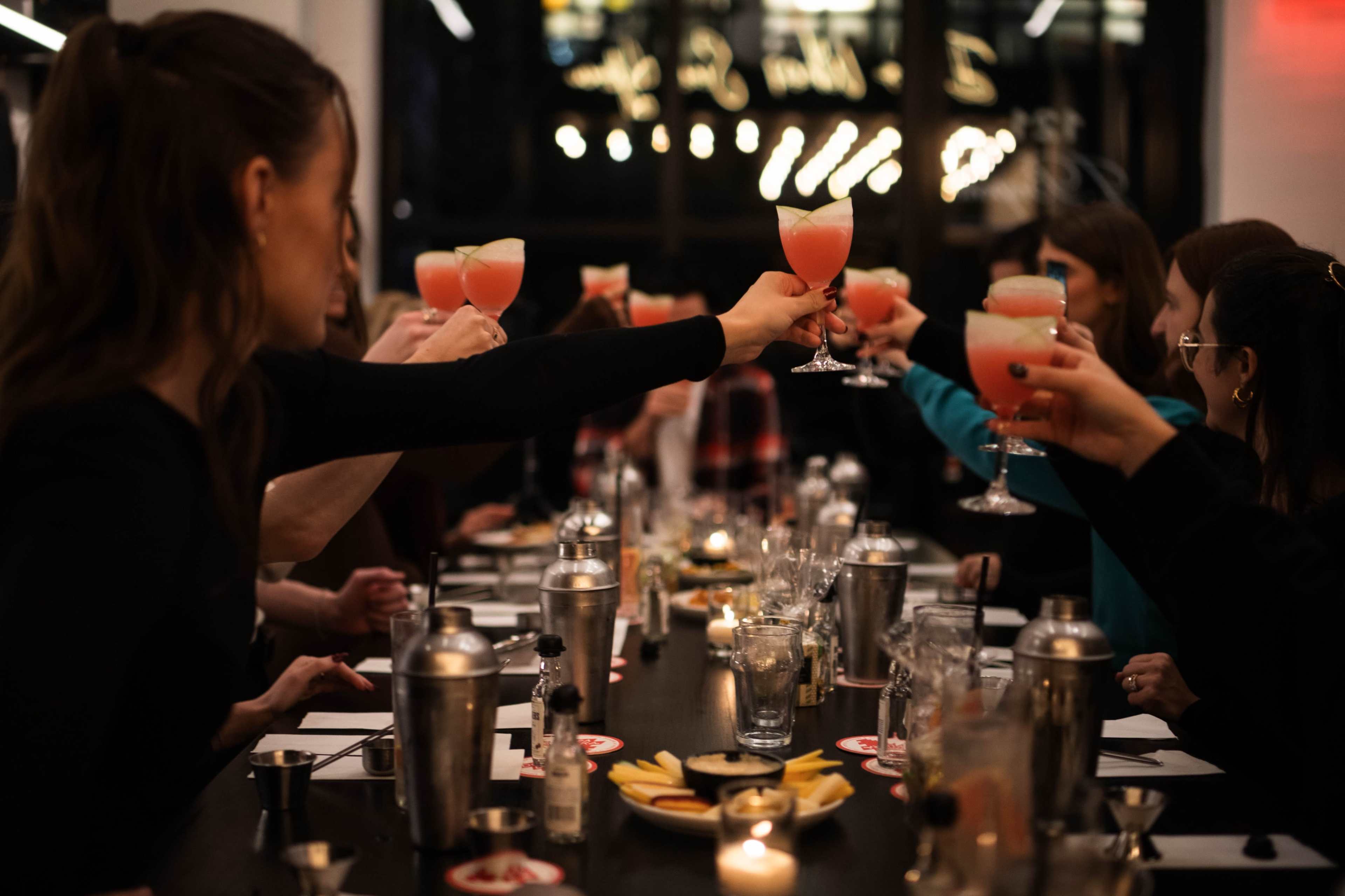 A group of people at a long table raise cocktail glasses in a toast during a social gathering.