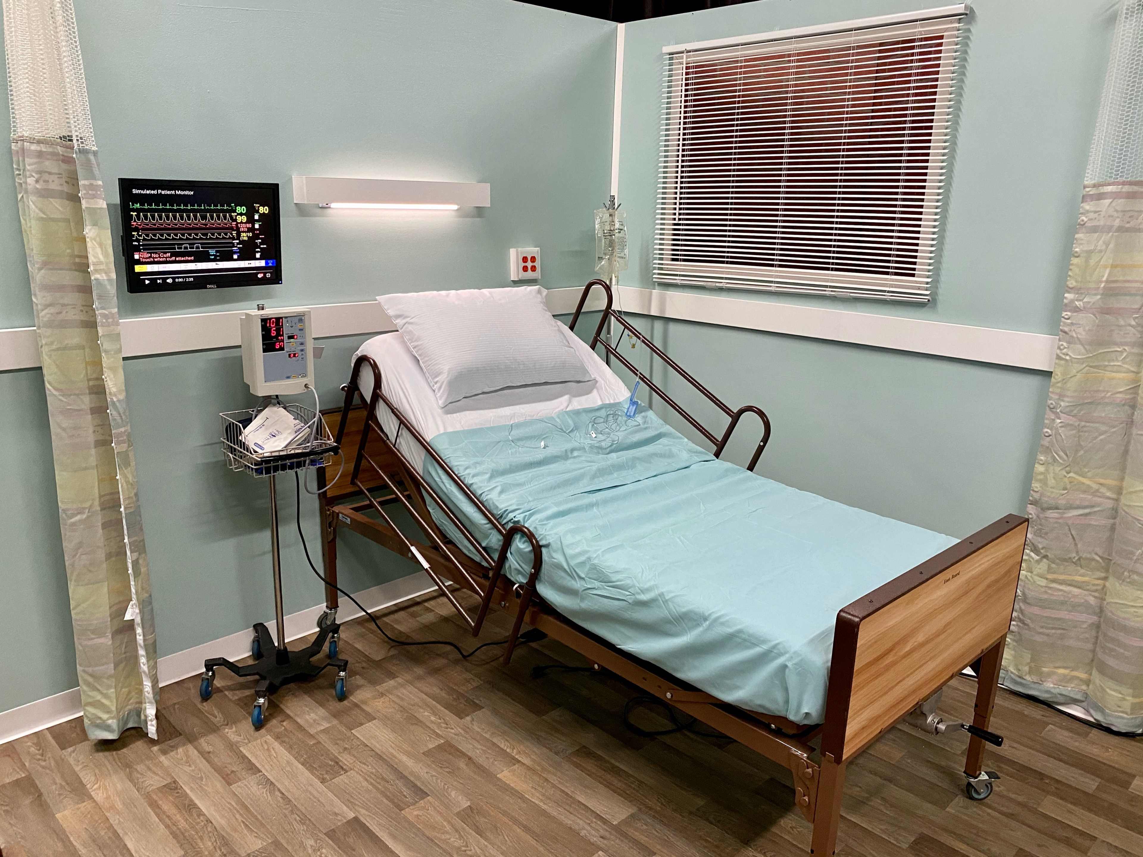 A hospital room featuring an adjustable bed with a light blue sheet, a monitor displaying medical data, and a small rolling cart with medical supplies.