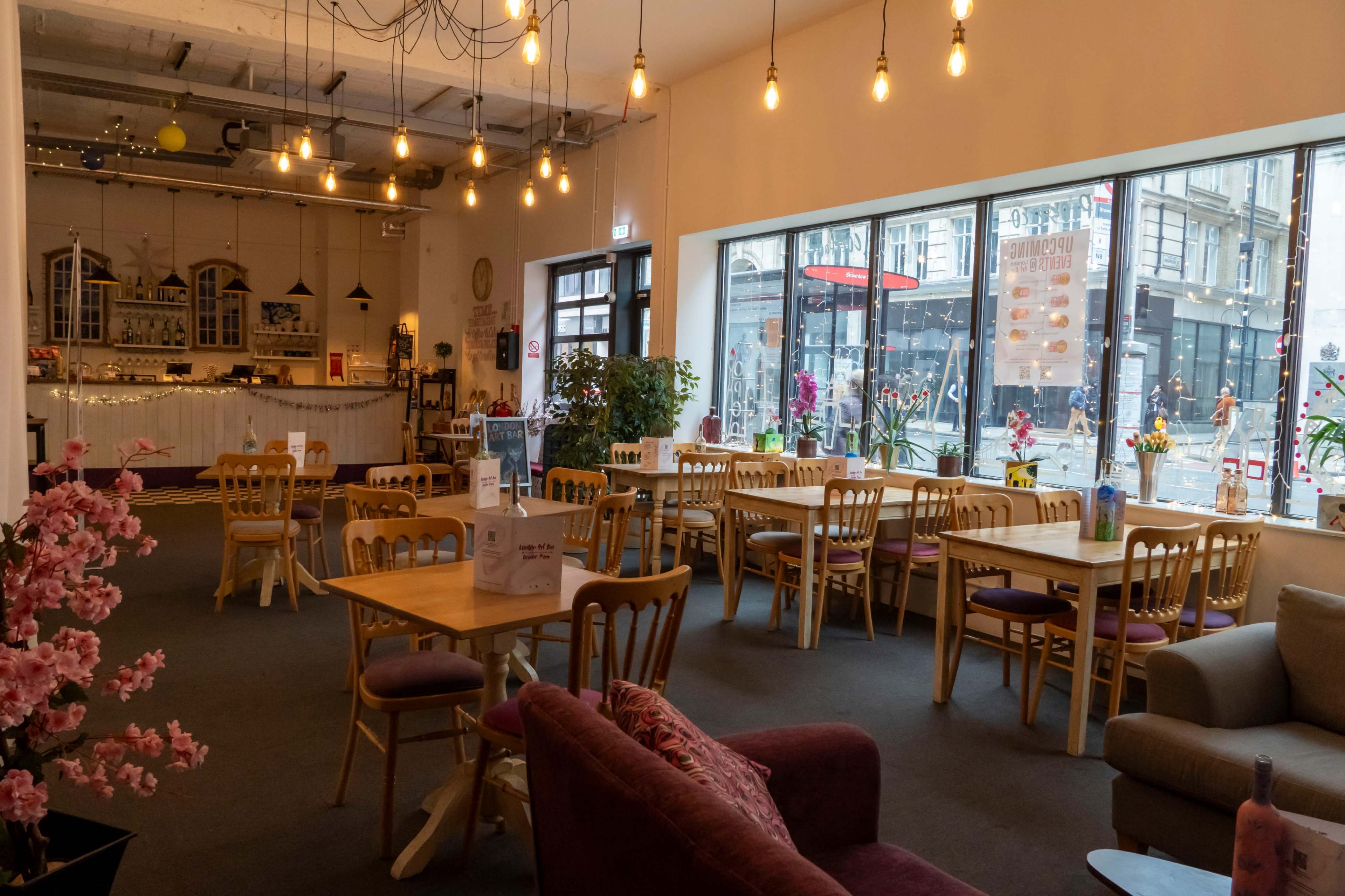 The image shows a cozy café interior with wooden tables, chairs, and large windows that overlook a bustling street.