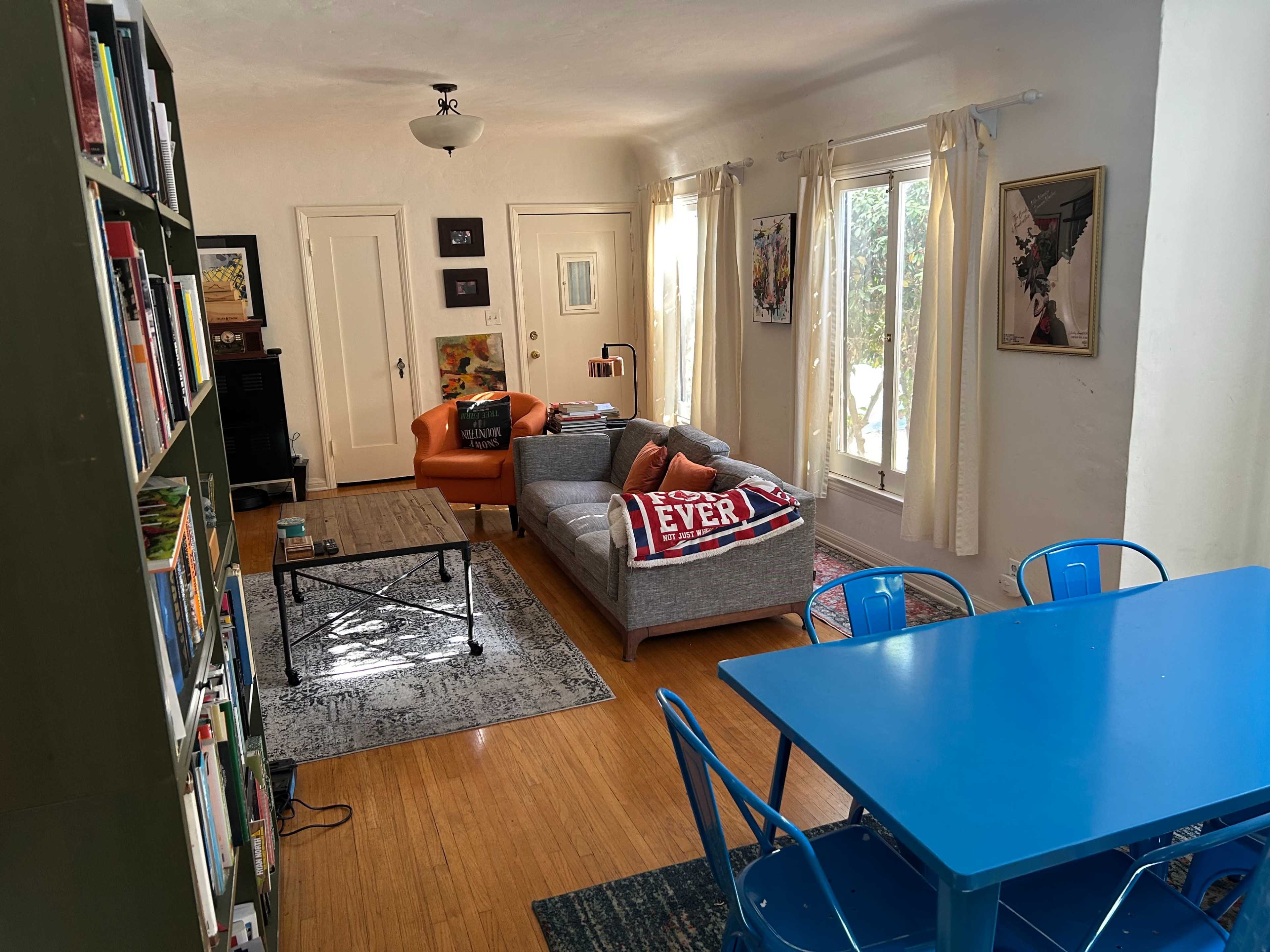 The image shows a cozy living space featuring a seating area with a gray couch and orange sofa, a coffee table, a blue dining table with chairs, and bookshelves lining one wall.
