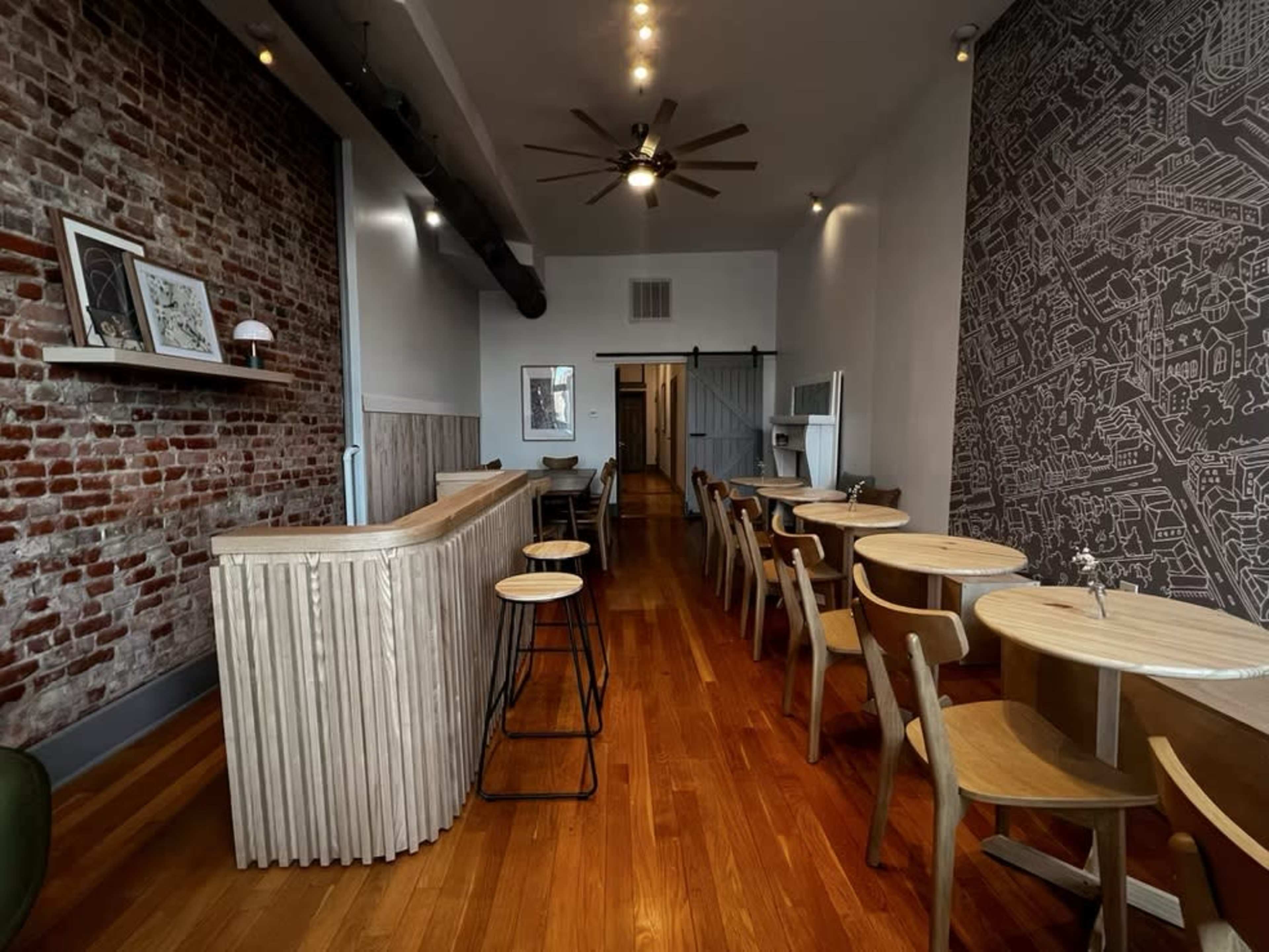 The image shows a modern cafe interior with exposed brick walls, wooden flooring, and a mix of round and rectangular tables arranged throughout the space.
