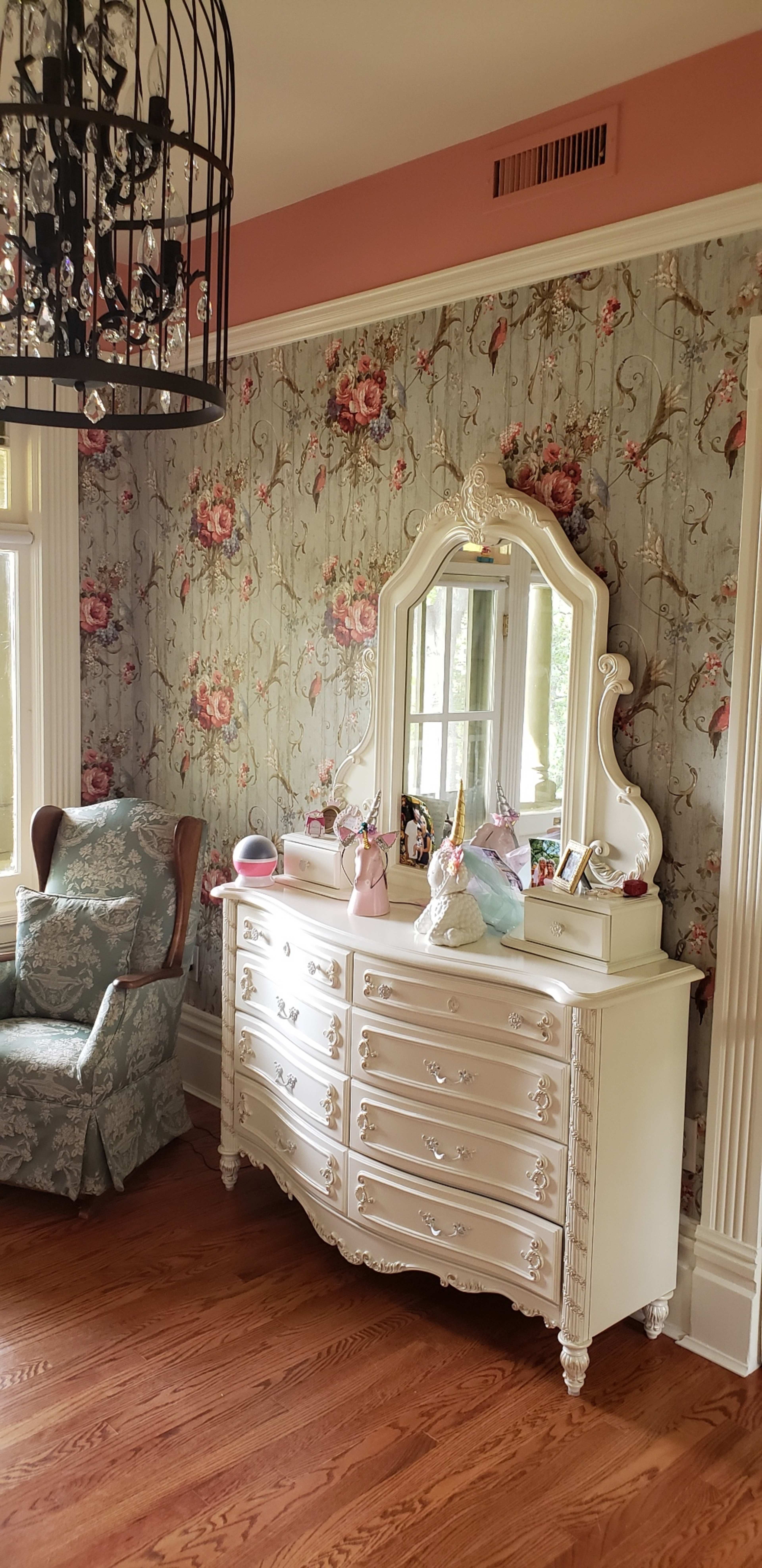 The room features a white dresser with a mirror, a patterned floral wall, and an armchair in the corner, all illuminated by a chandelier.