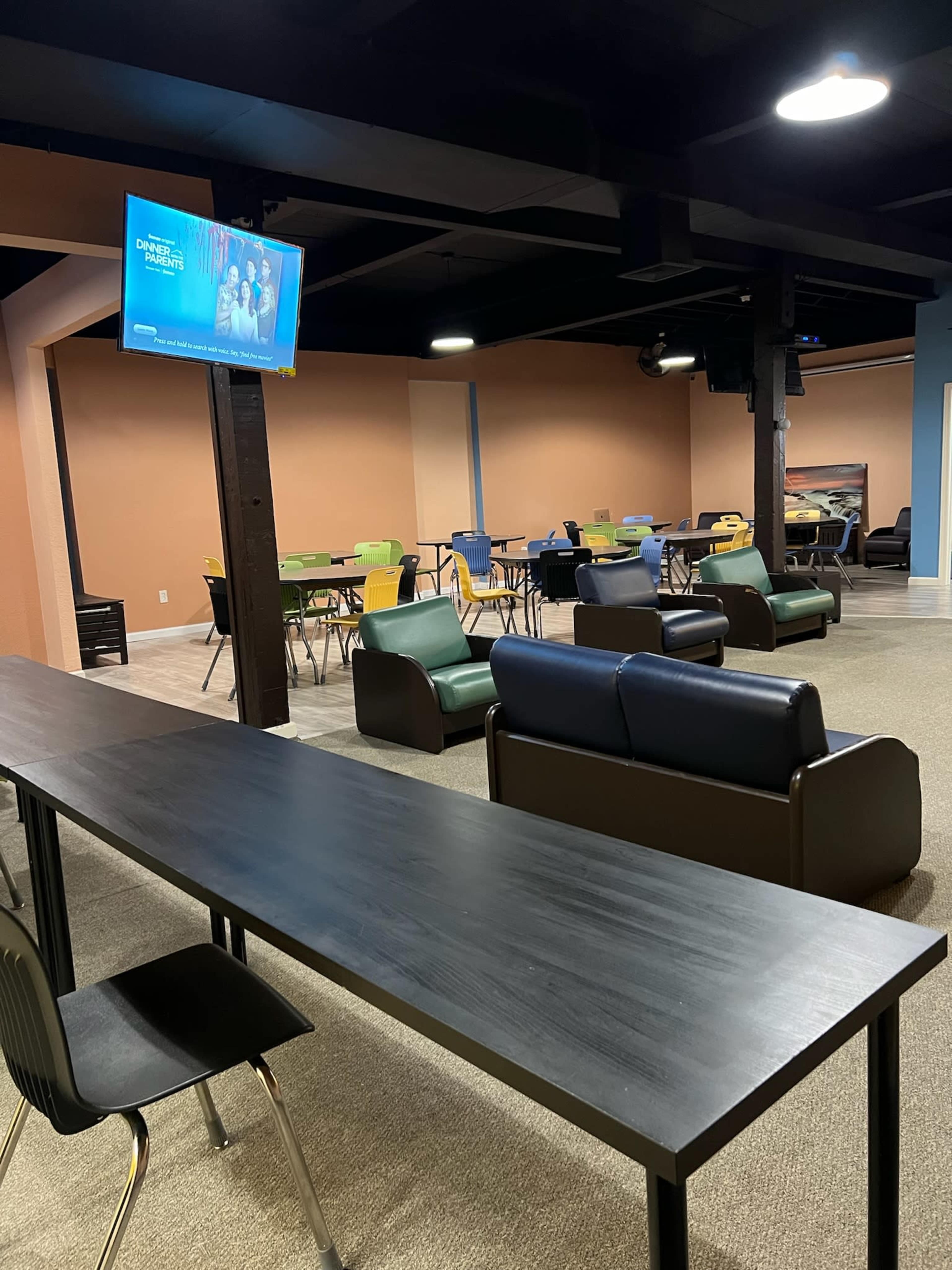 The image shows a spacious communal area with various seating arrangements, including tables and chairs, along with a television mounted on the wall.