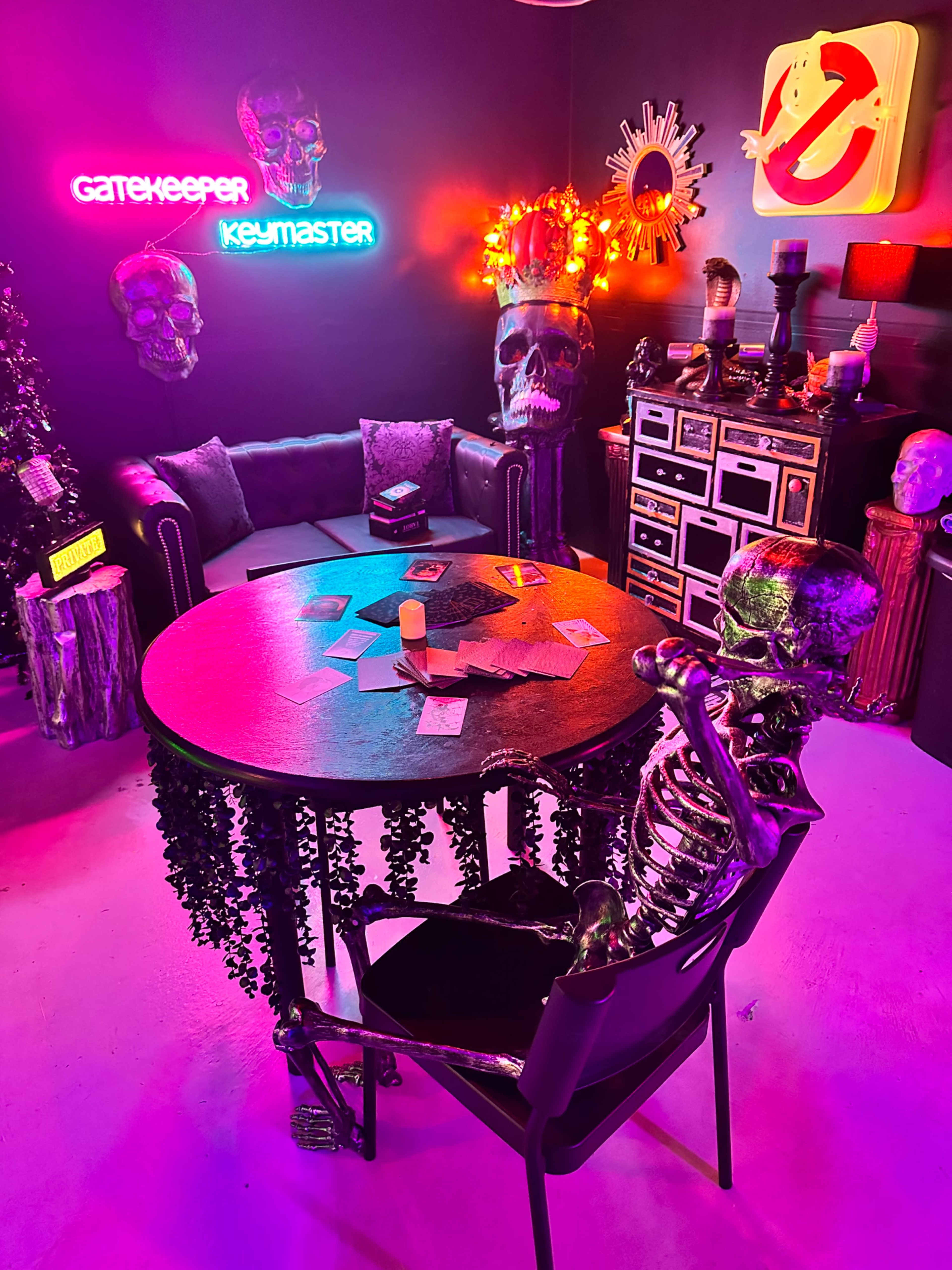 The image shows a uniquely decorated room featuring a round table with a skeleton seated at it, vibrant neon signs, and various spooky decor elements including skulls and plants.