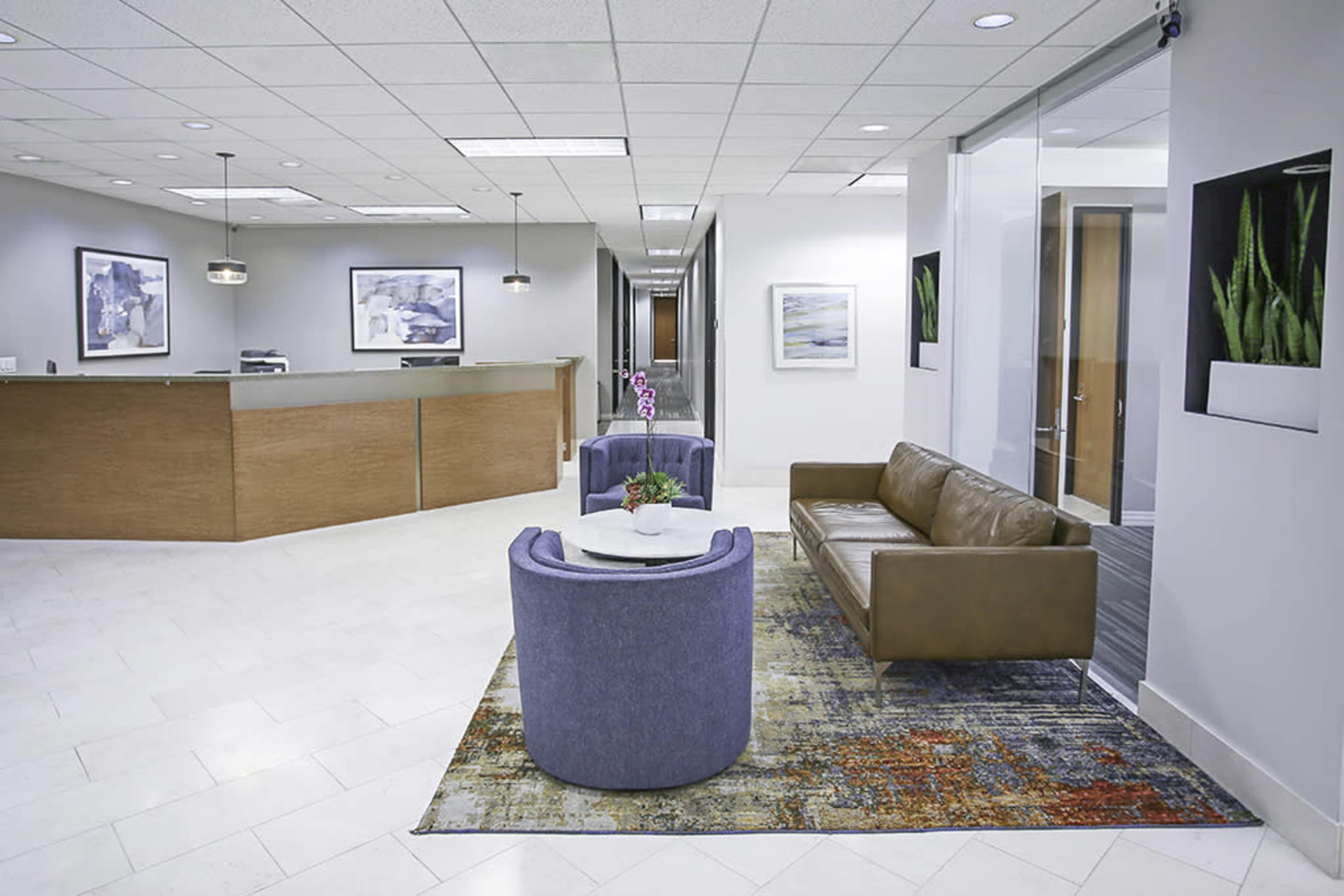 A modern office reception area featuring a waiting room with two upholstered chairs, a leather sofa, a round table, and artworks on the walls.