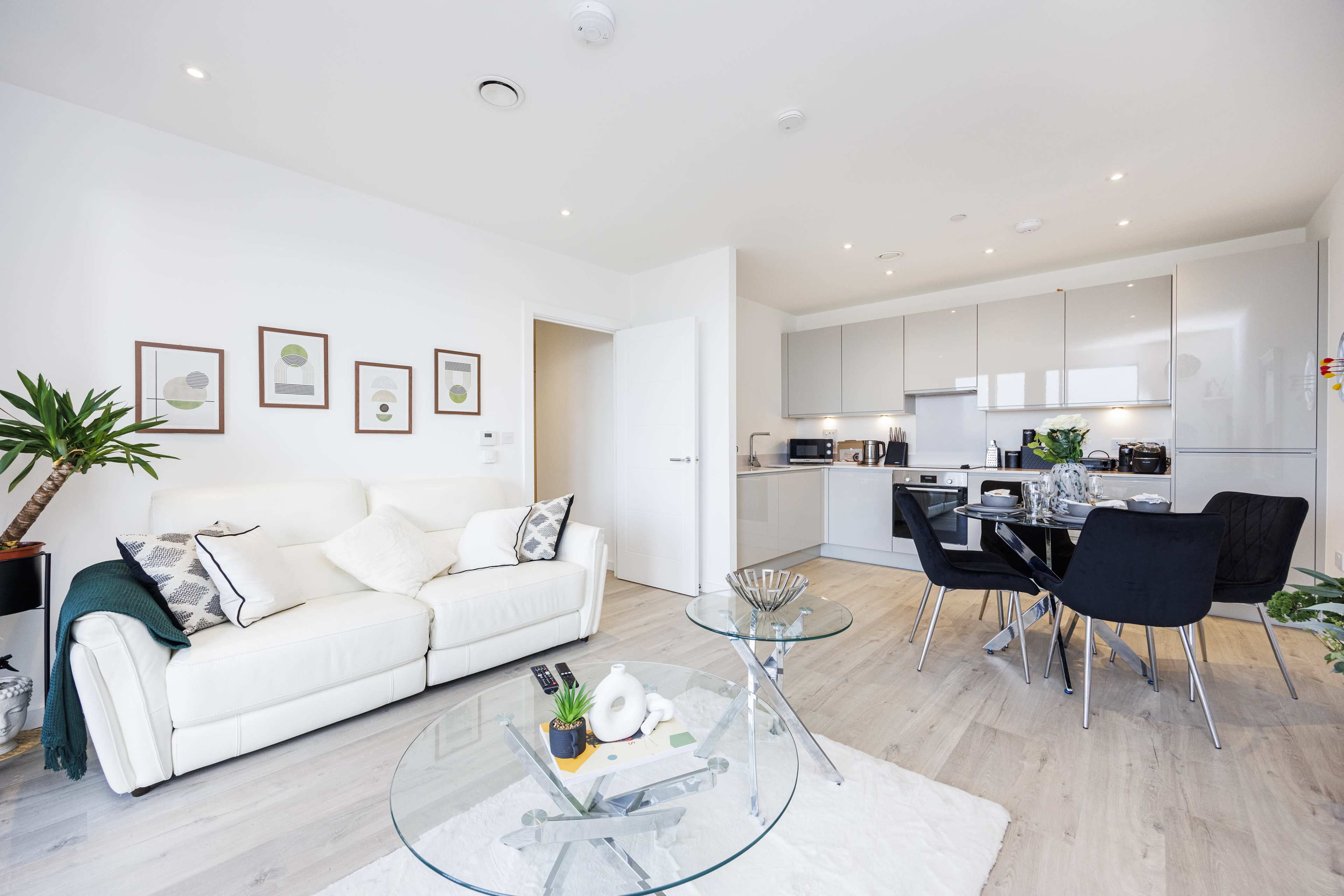 A modern living room with a white sofa and a glass coffee table, leading into a sleek kitchen with gray cabinets and stainless steel appliances.