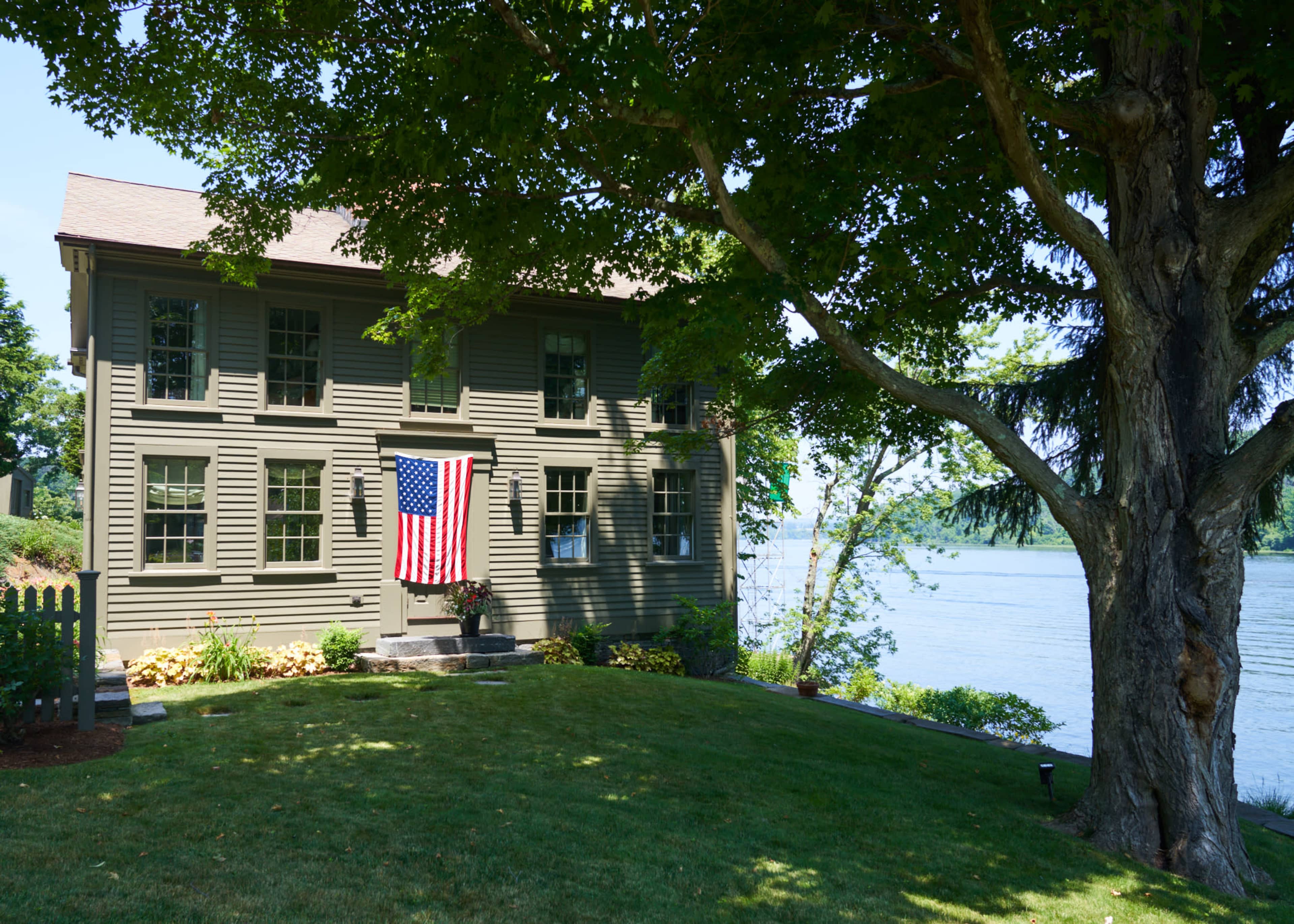 A two-story green house with an American flag hangs on its front, situated next to a river and surrounded by trees.