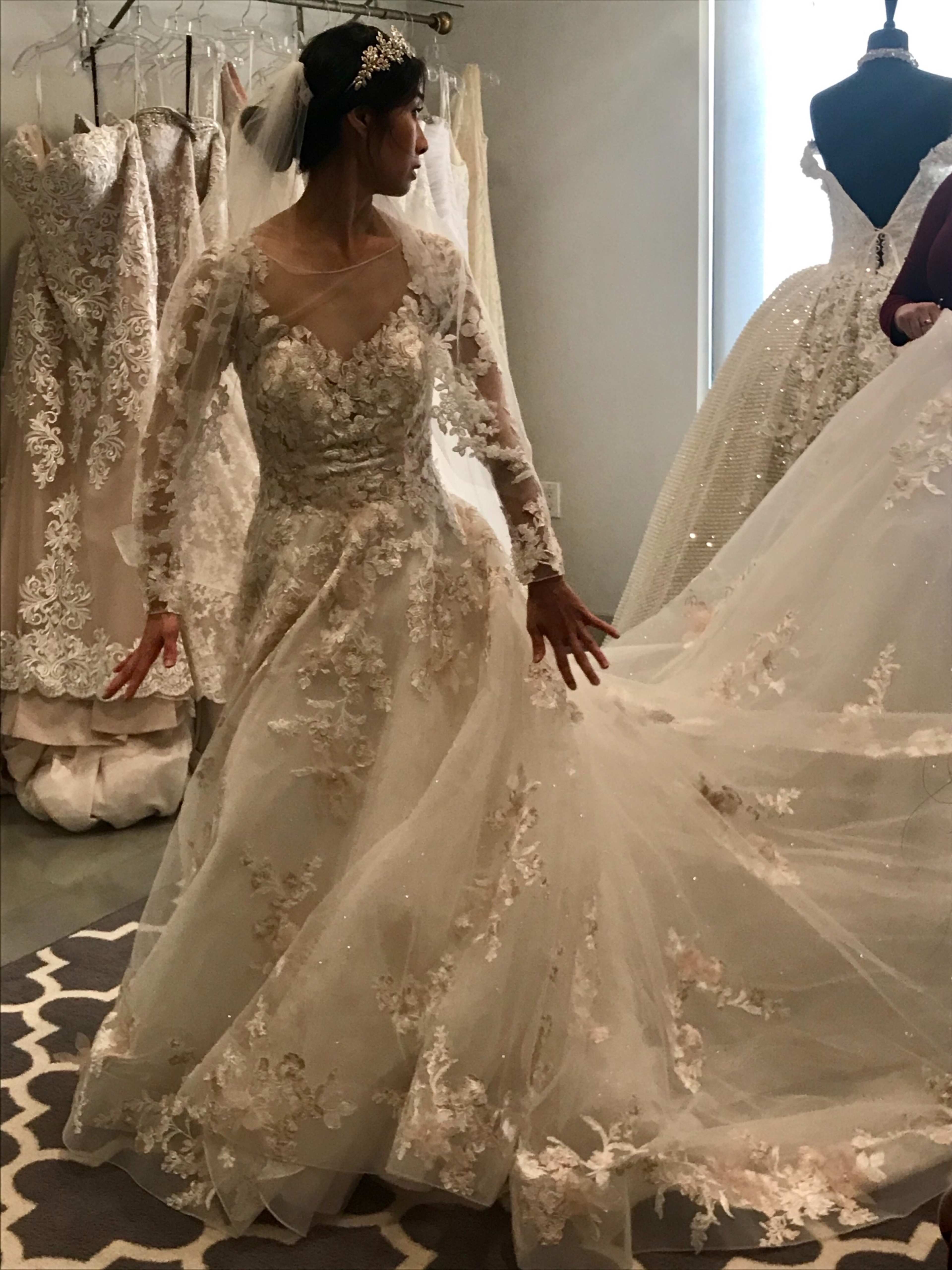A woman is trying on an intricately designed wedding dress in a boutique, surrounded by other gowns on display.
