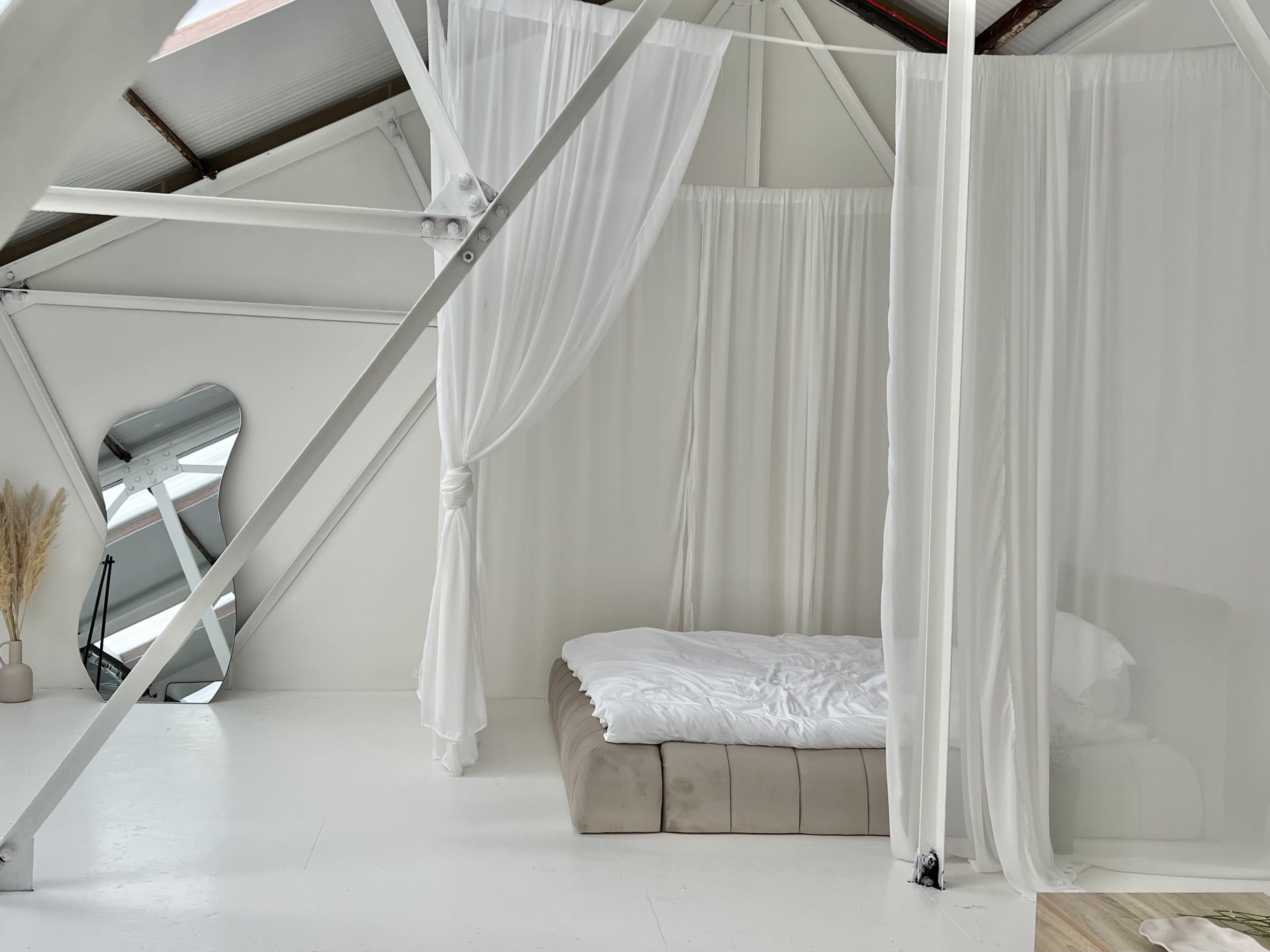A minimalist bedroom featuring a bed with white linens, surrounded by sheer curtains and a mirror against a white wall.
