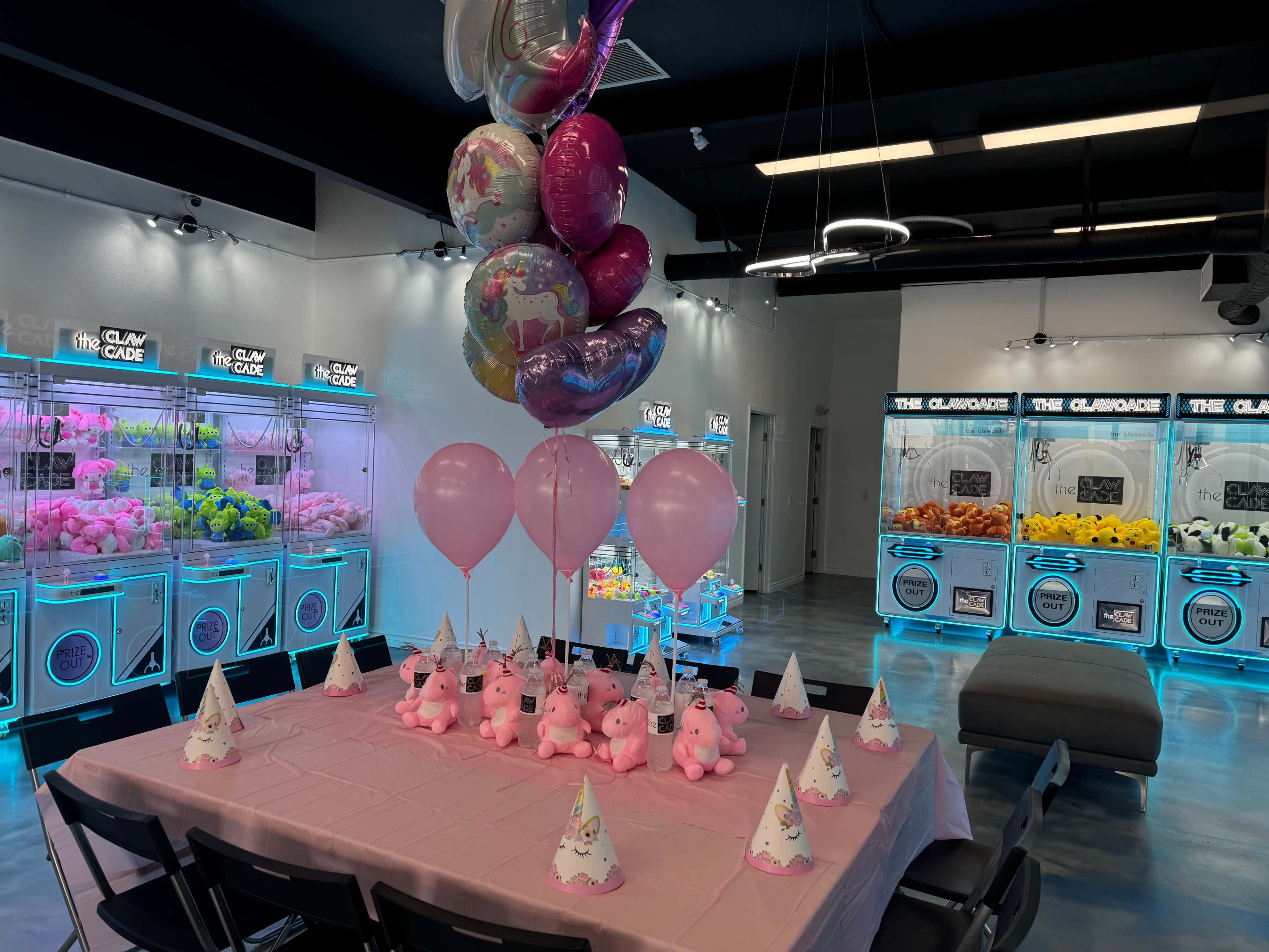 The image shows a party setup with a table adorned with pink balloons and plush pig centerpieces, surrounded by arcade machines in a brightly lit room.