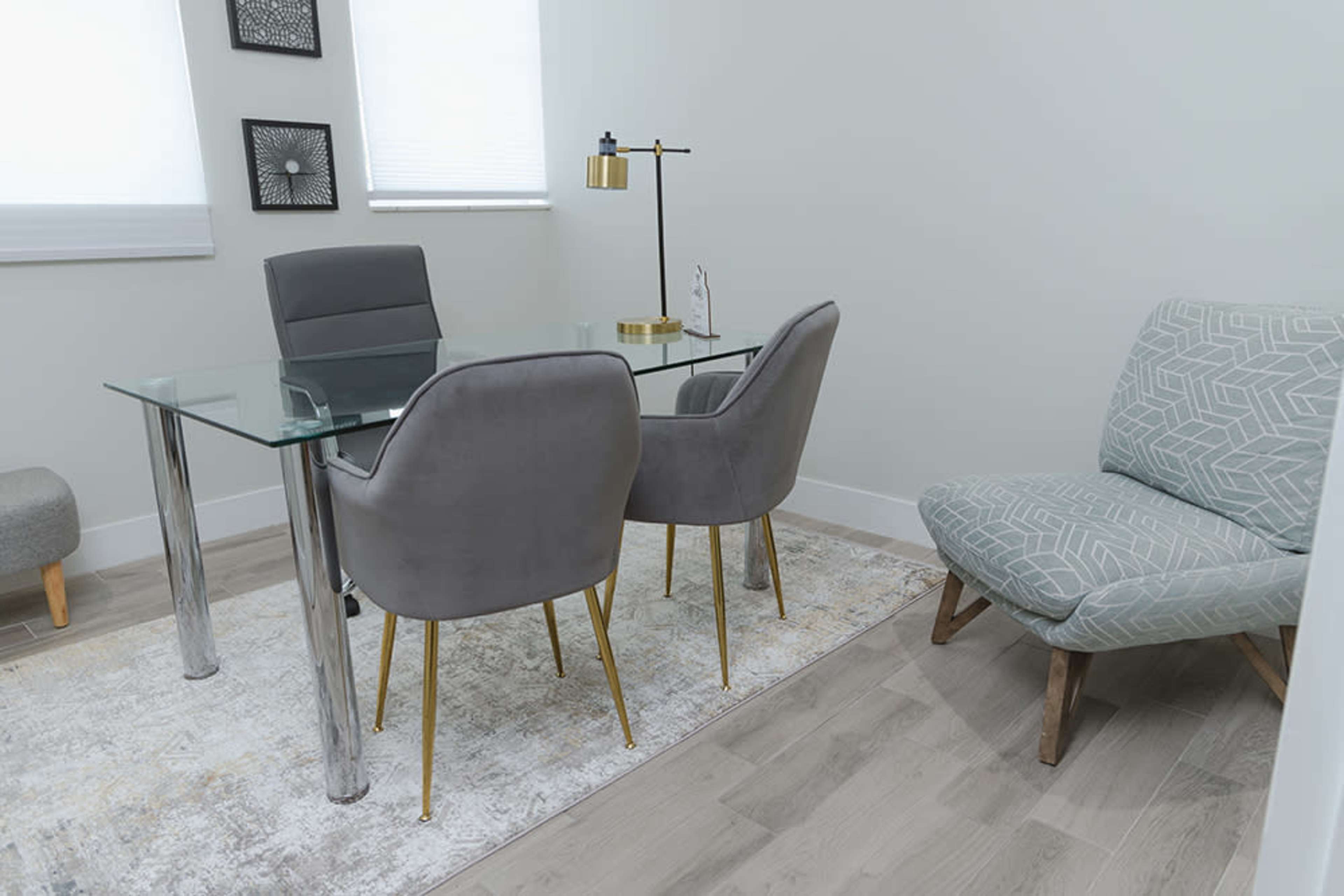 A modern office space features a glass-topped table with two gray chairs and a patterned armchair in the corner.