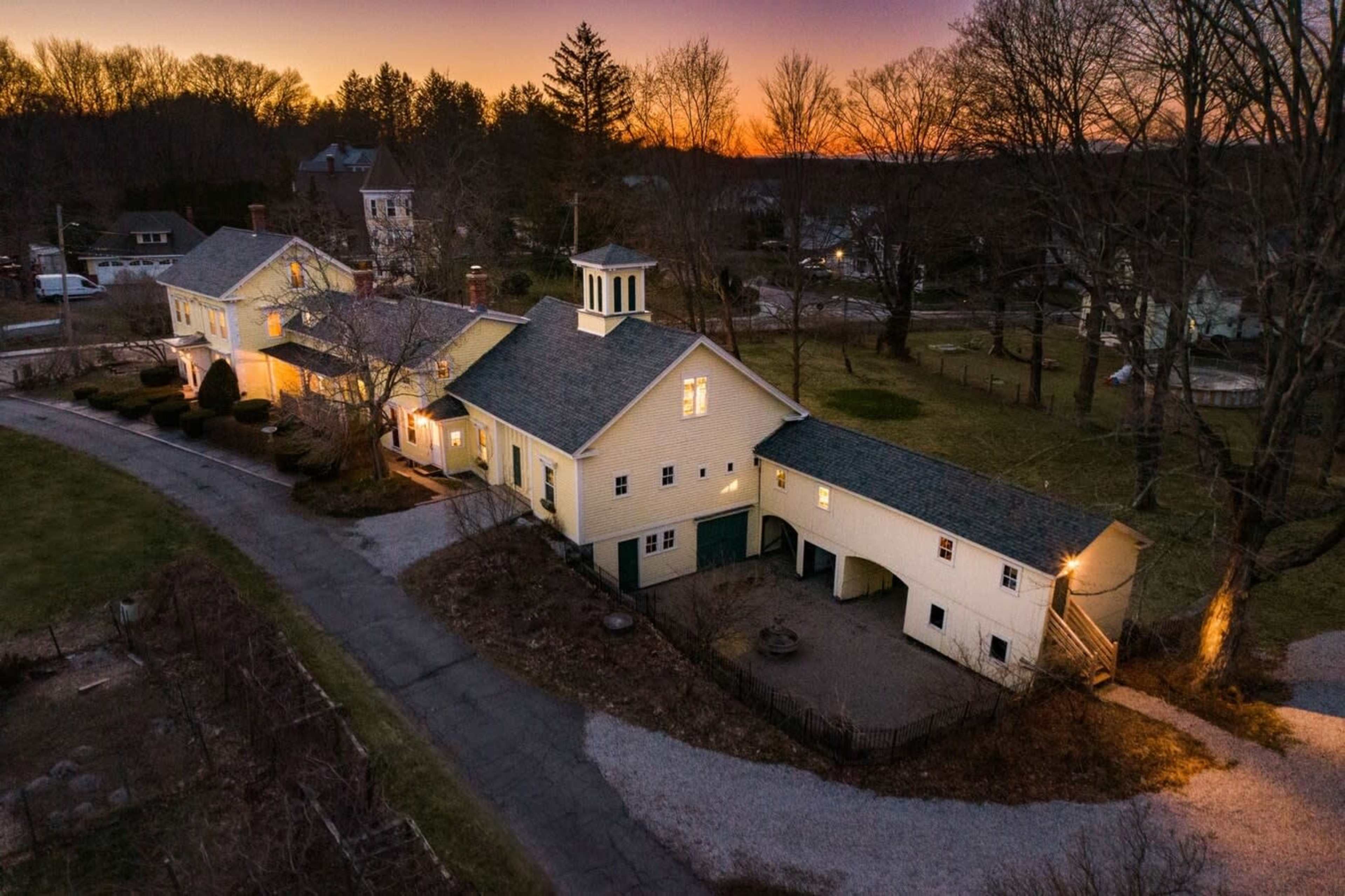 A large, two-story yellow house with a cupola is situated at the end of a gravel driveway, surrounded by trees during sunset.