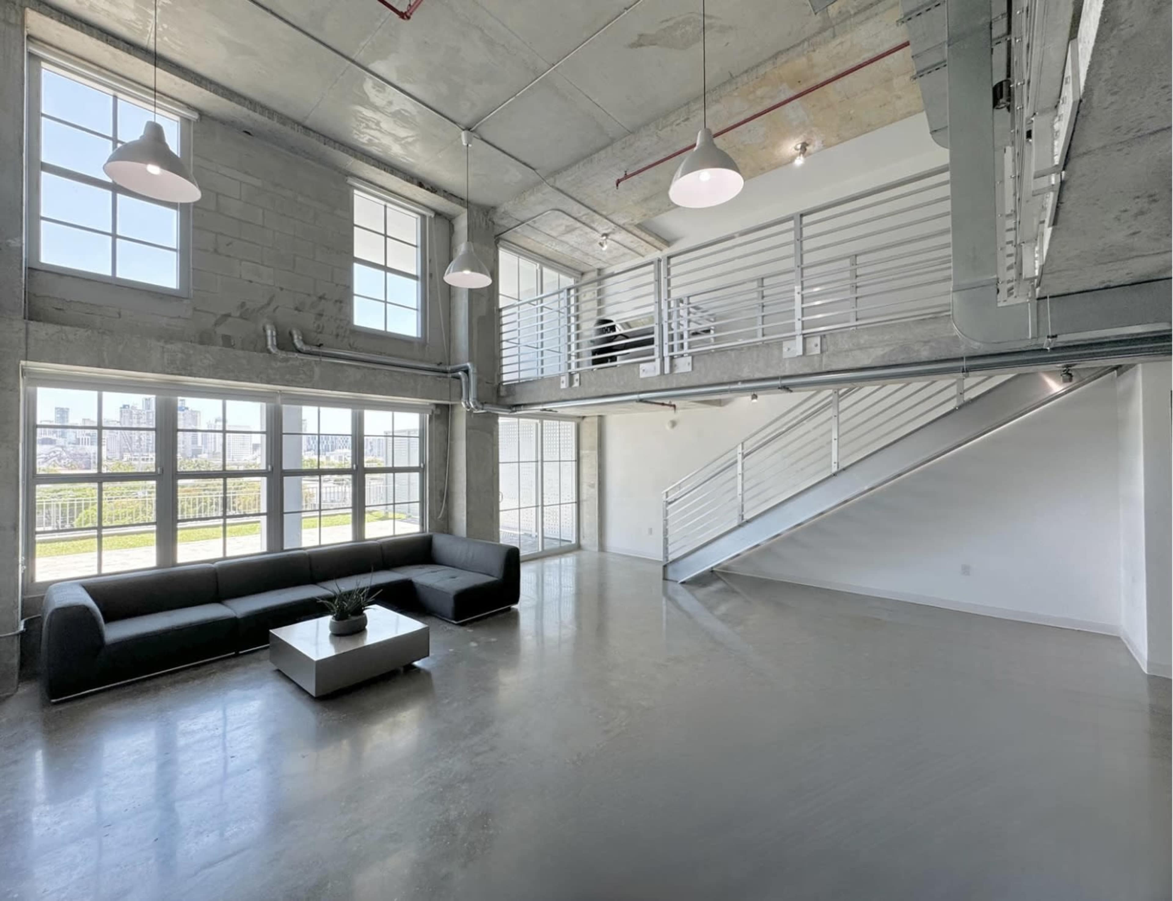 The image shows a modern, open-concept living space with large windows, a gray couch, a coffee table, and a staircase leading to an upper level.