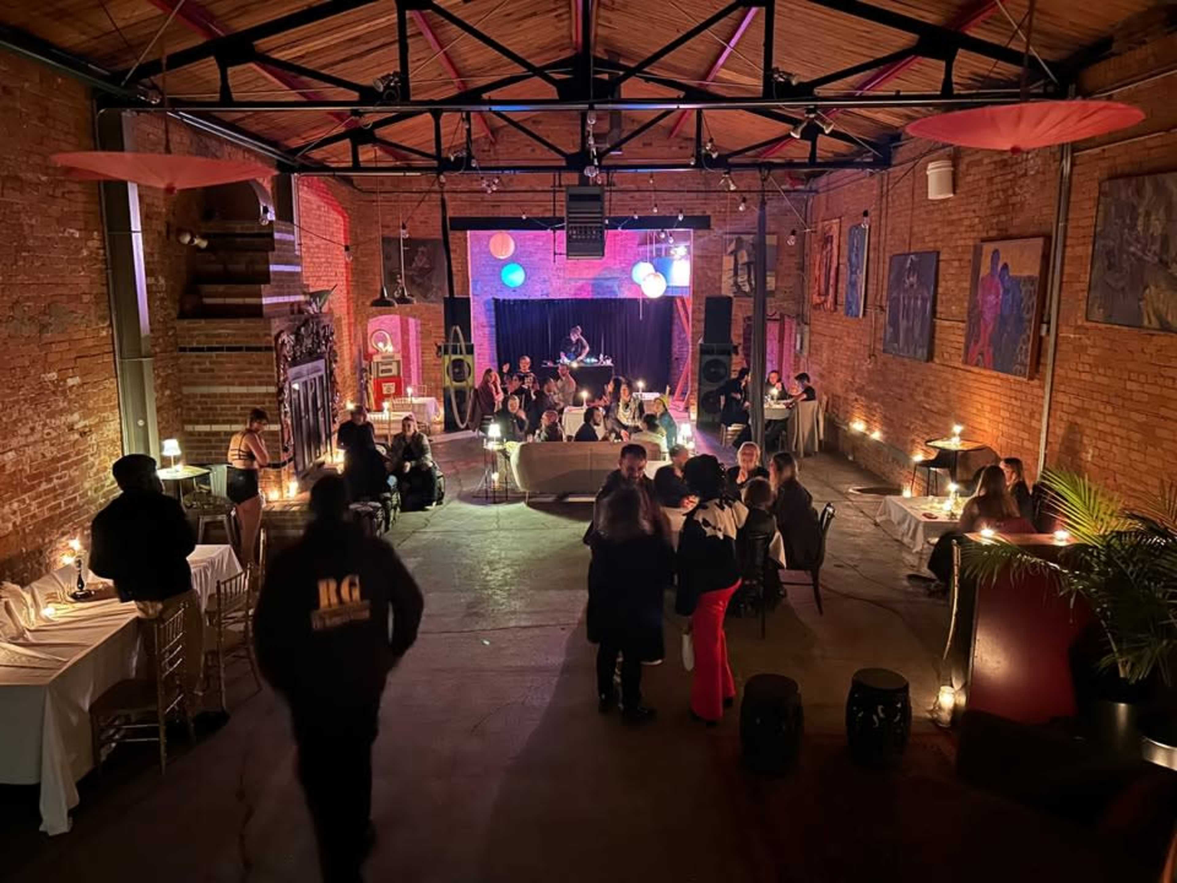 A crowded event space with brick walls features tables, attendees mingling, and a stage with colorful lighting at one end.