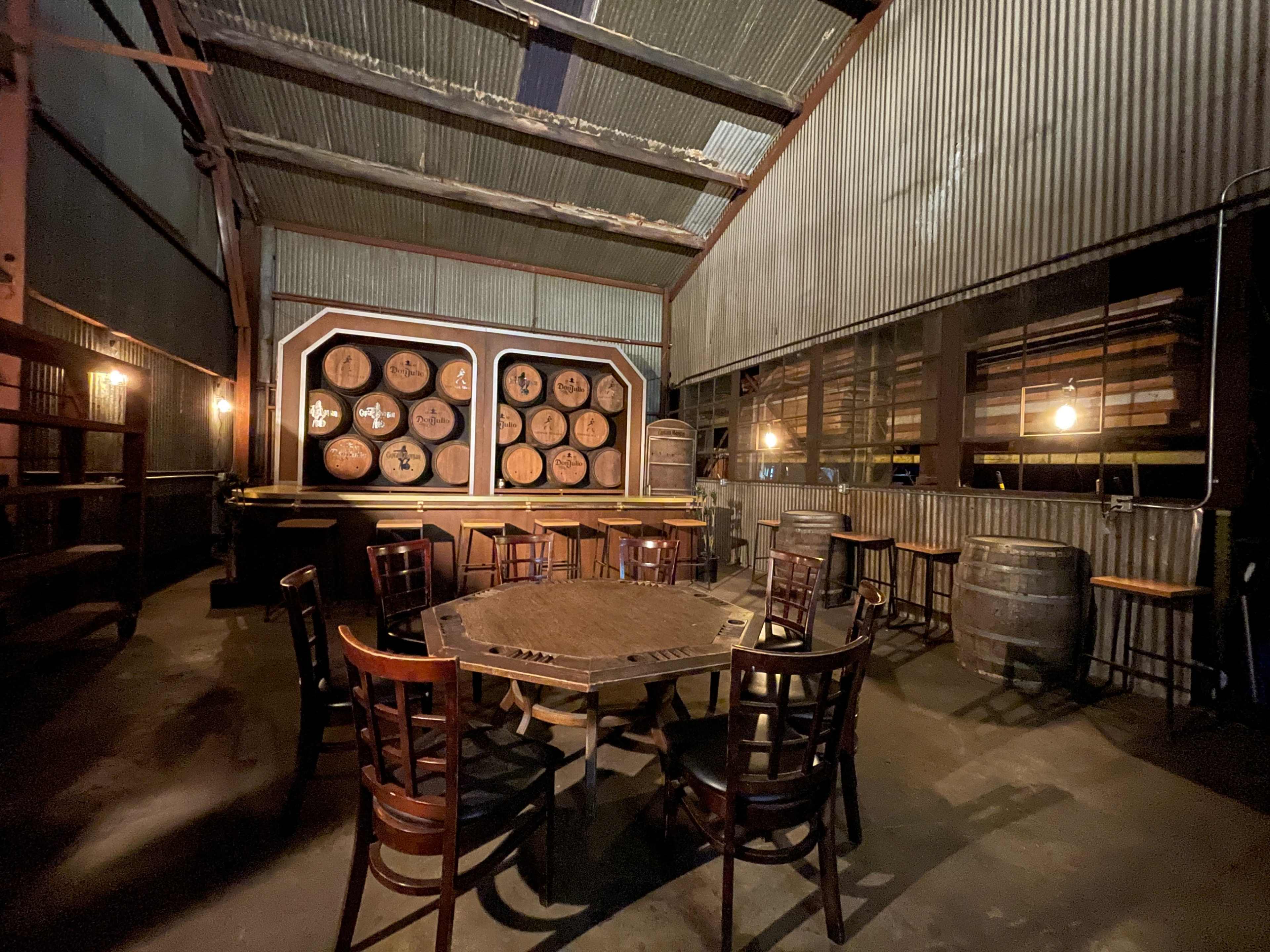 The interior features a dimly lit room with wooden barrels and a set of circular wine barrels mounted on the wall, accompanied by a round table and several wooden chairs.