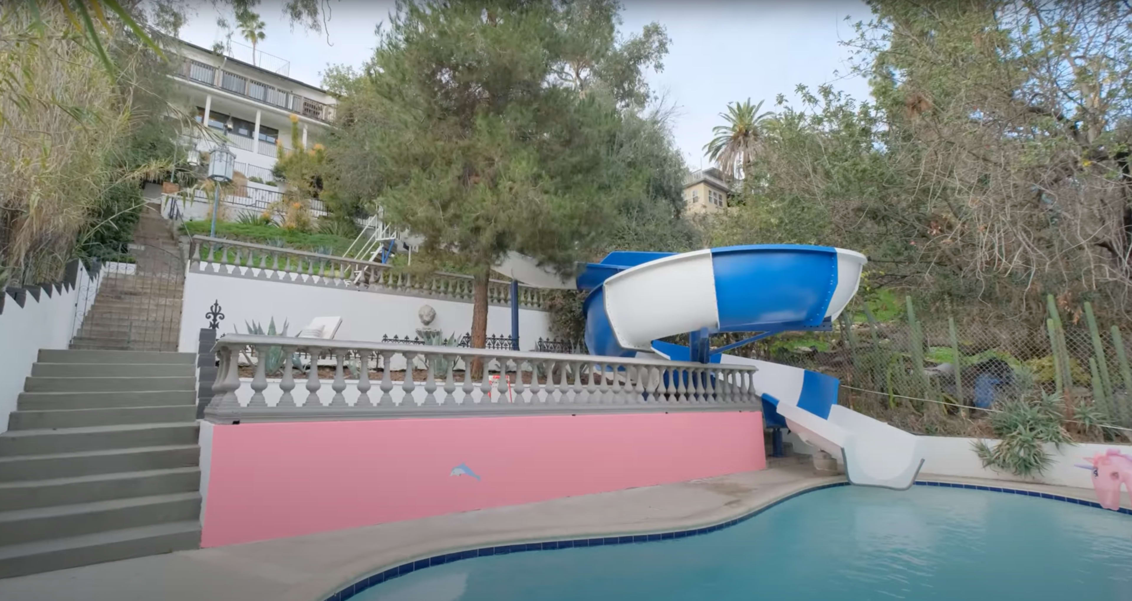 A swimming pool with a large blue water slide next to a set of stairs leading to a hillside property.