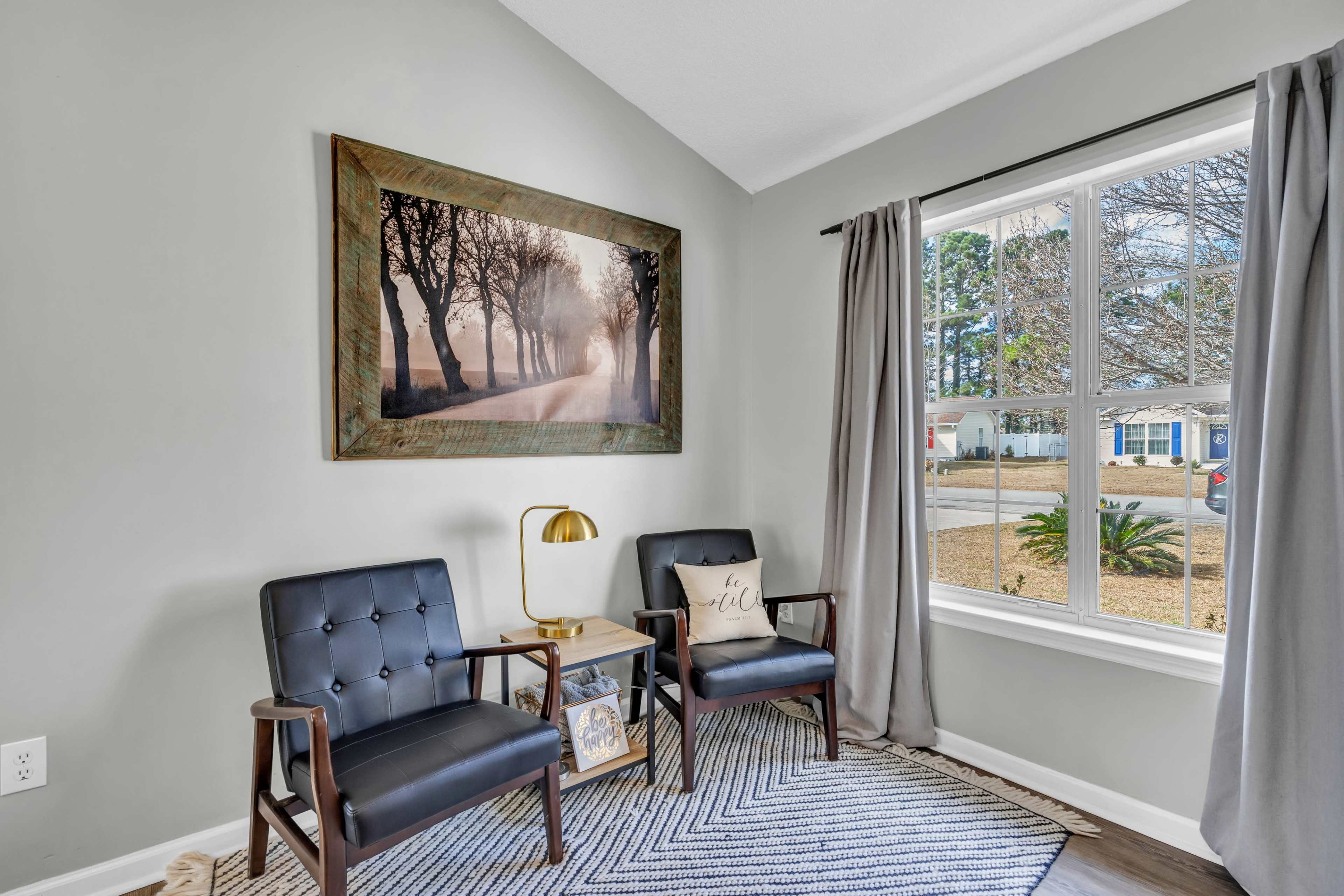 A cozy corner of a room with two black chairs, a small side table, and a large framed picture hanging on the light green wall, with a window displaying a view of a front yard.